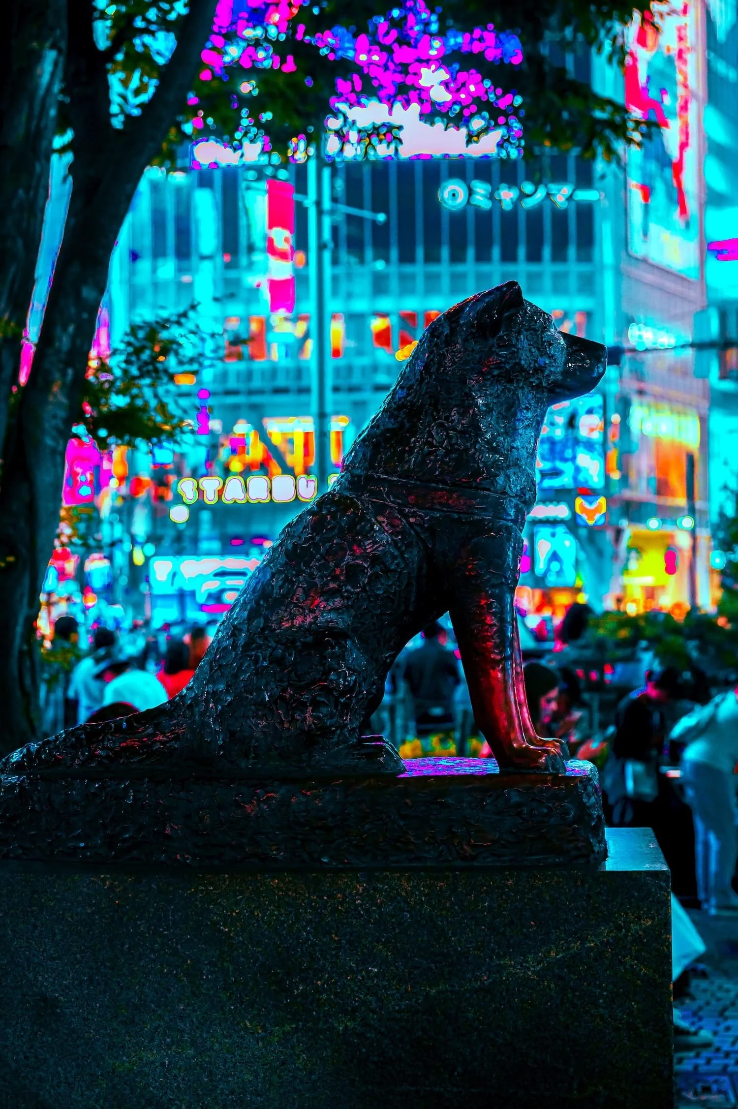A black dog statue on a pedestal in a lively, neon-lit urban street scene at night, with people and colorful signs in the background.