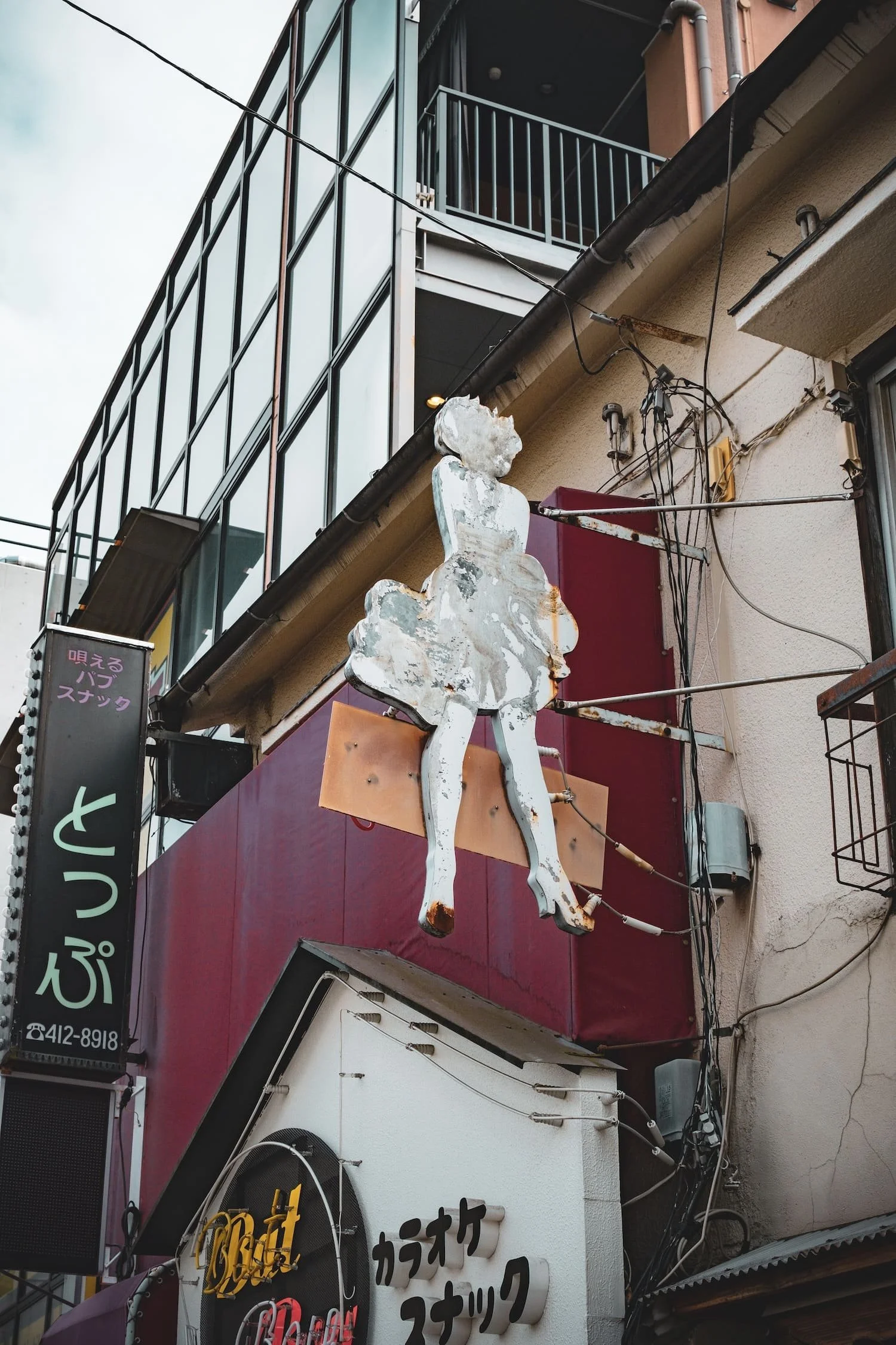 A vintage, worn-out metallic sign of a woman sitting, attached to a building with electrical wires around.