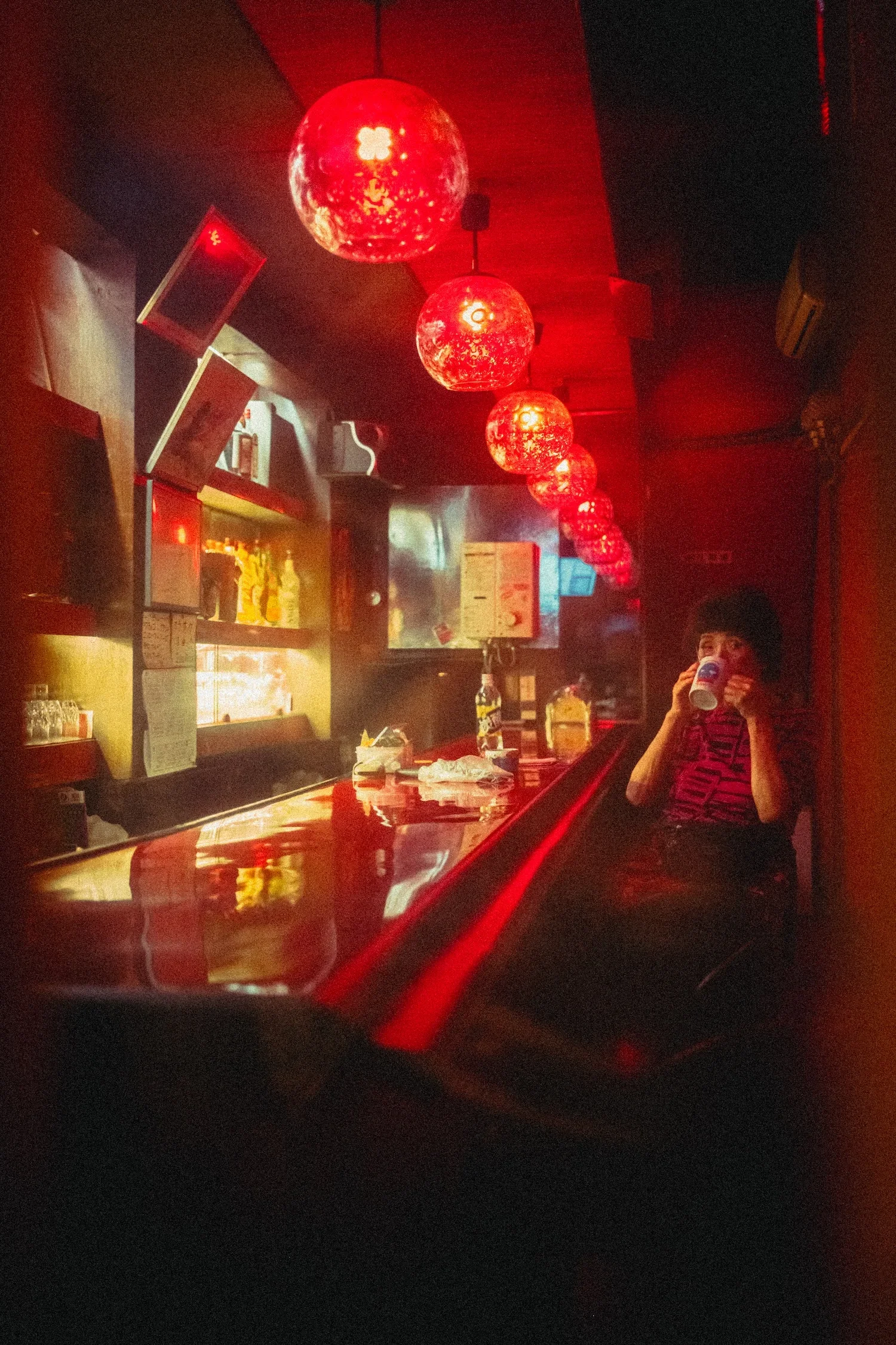 A person with dark hair holding a drink in a dimly lit bar, illuminated by red hanging lights.