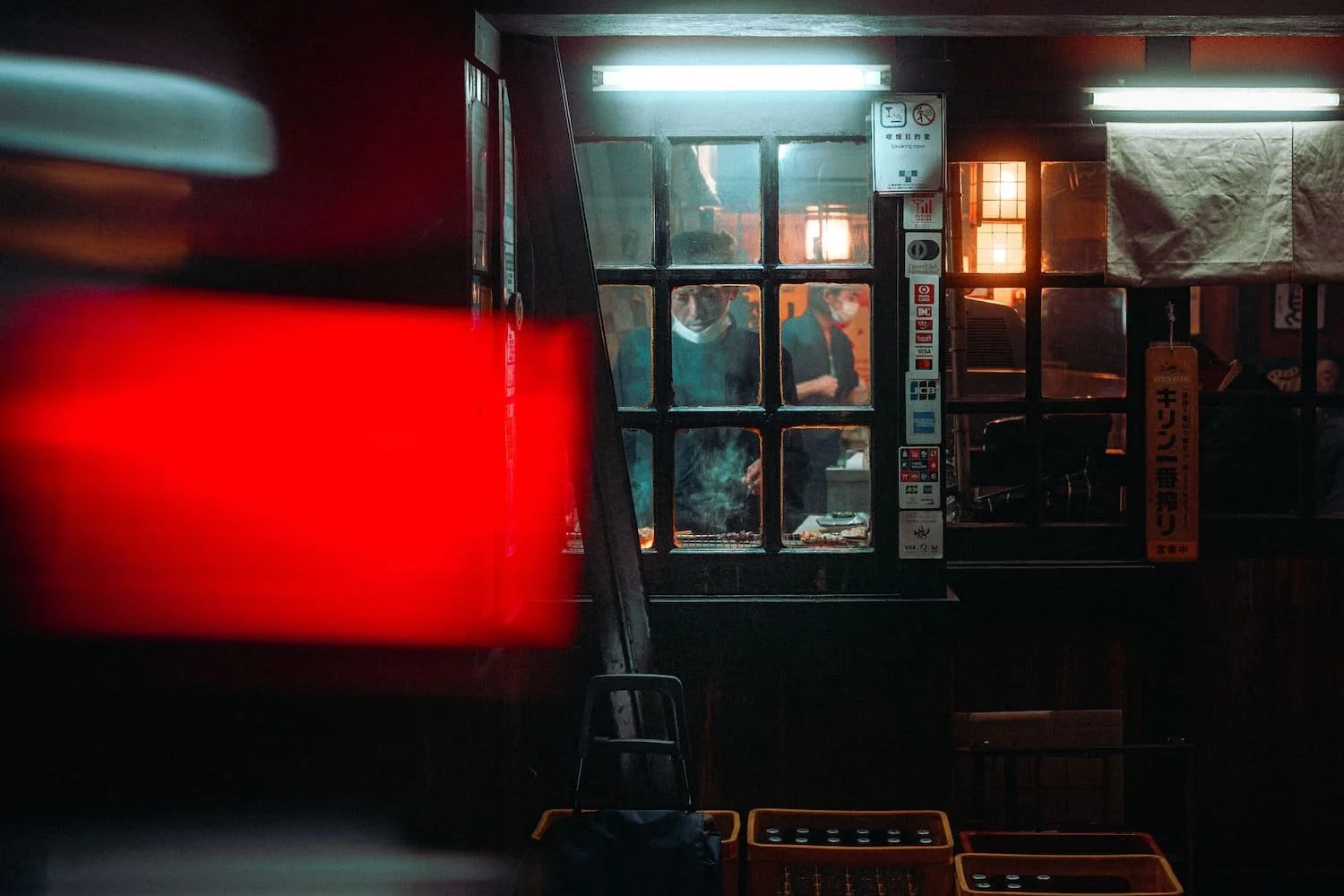 People inside a dimly lit restaurant seen through a window with bars, with a red light in the foreground and various signs on the wall, some wearing masks.
