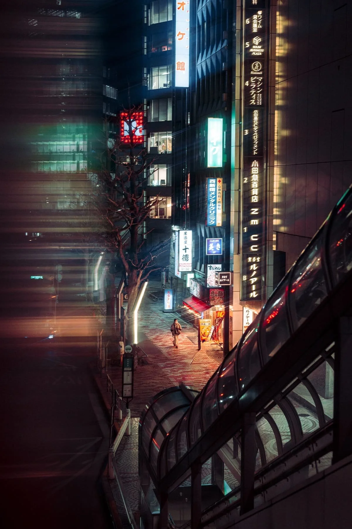 Night scene of a city street in Japan with bright neon signs, a leafless tree, a person walking, and an escalator in the foreground.