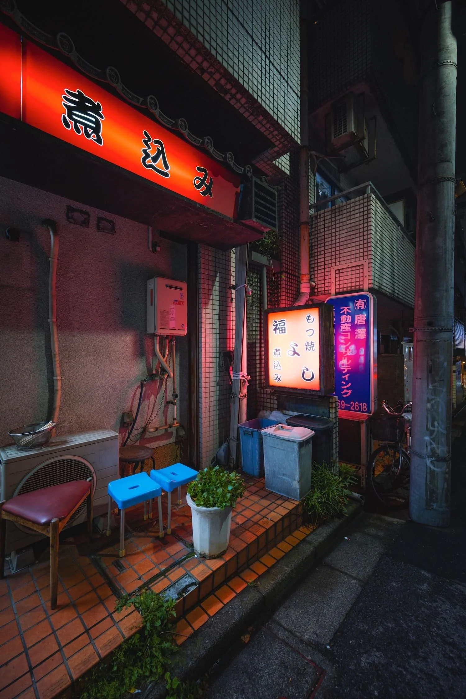 Night scene of a Japanese restaurant with illuminated signs, a bicycle, potted plants, and outdoor seating.