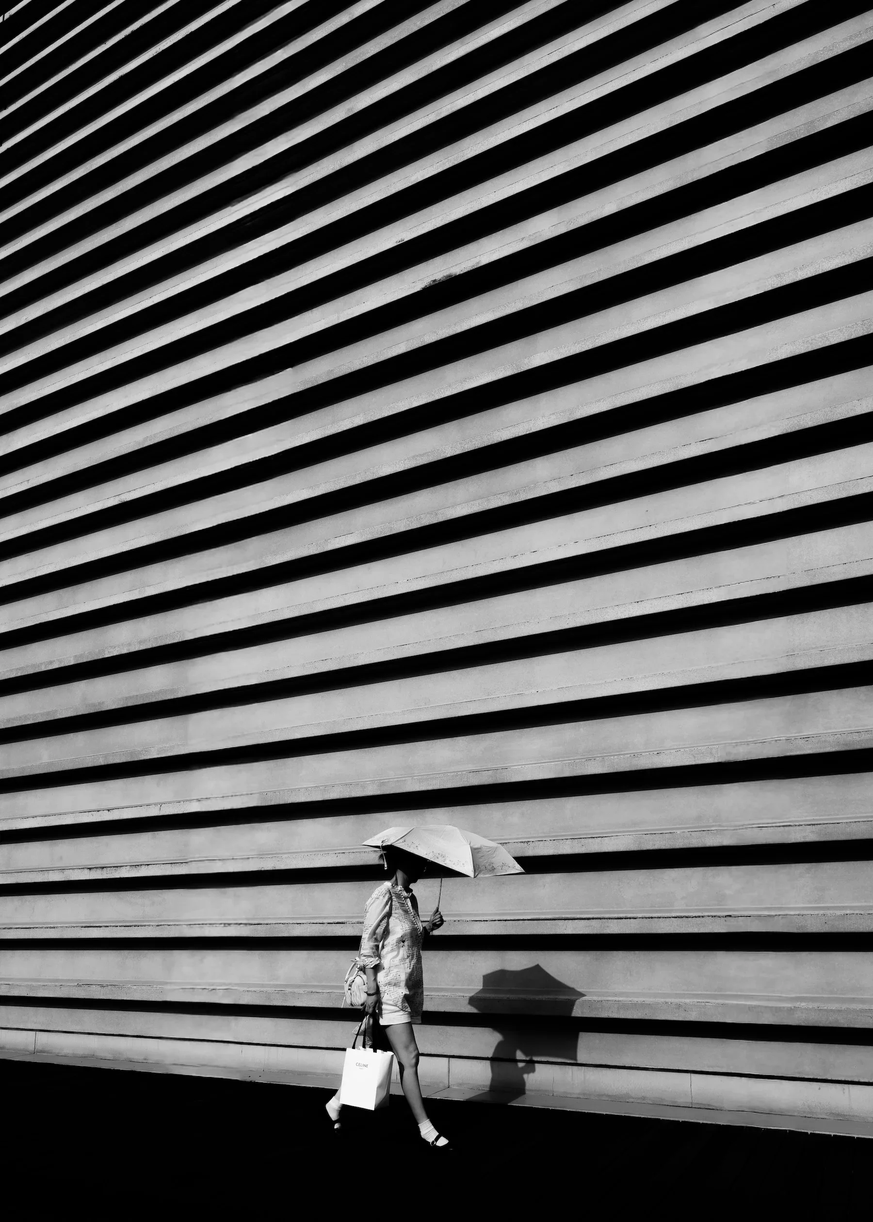 A woman holding an umbrella and a shopping bag with the word 'CELINE' on it, walking past a modern building with horizontal lines on its exterior in black and white.