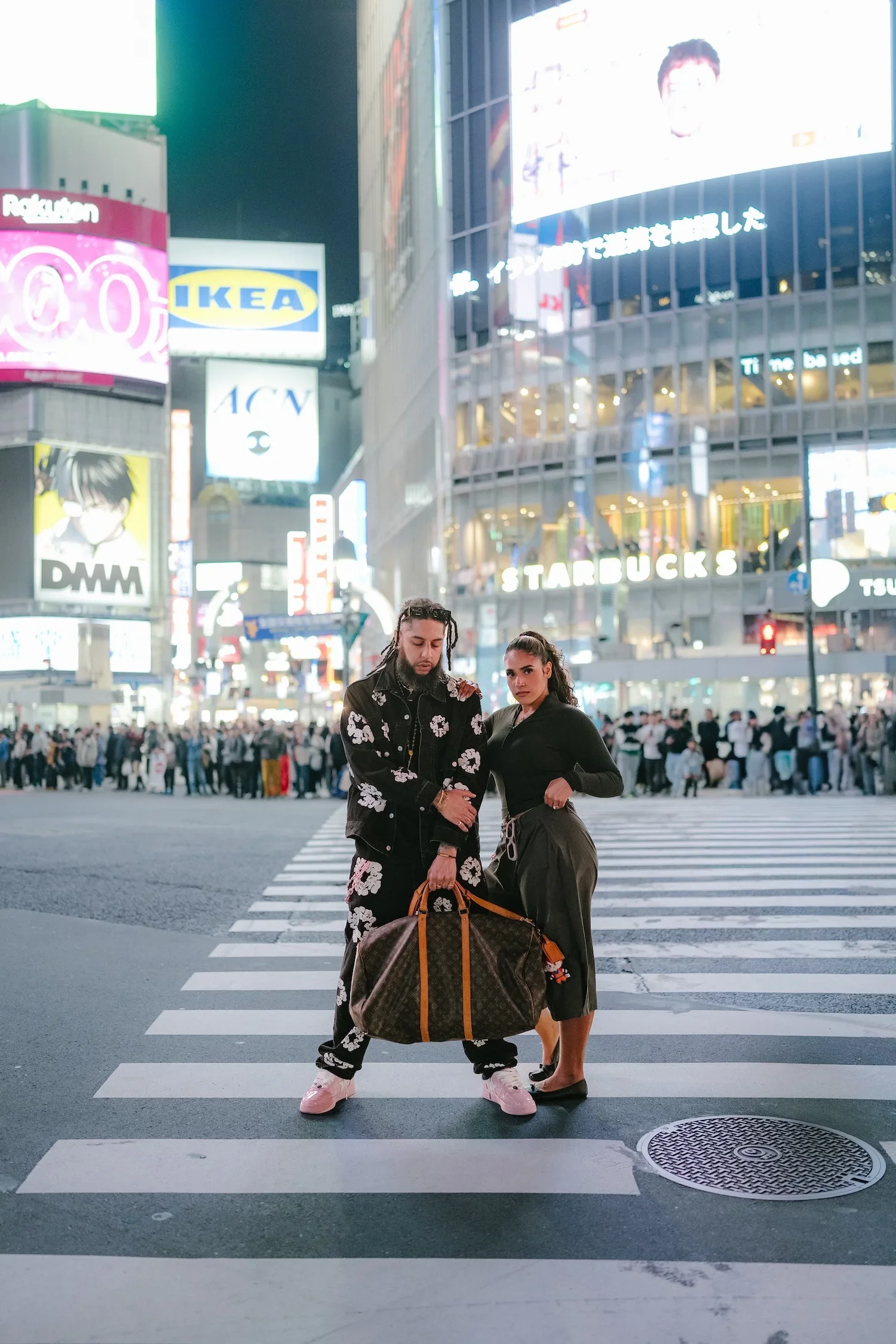 Two fashionably dressed people standing at a crosswalk in a busy city at night, surrounded by bright billboards and illuminated signs.