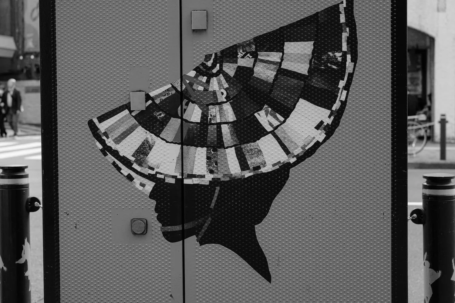 A metal city utility box featuring a black and white artwork of a hand holding a clock that resembles a fish, with various city streets and buildings in the background.
