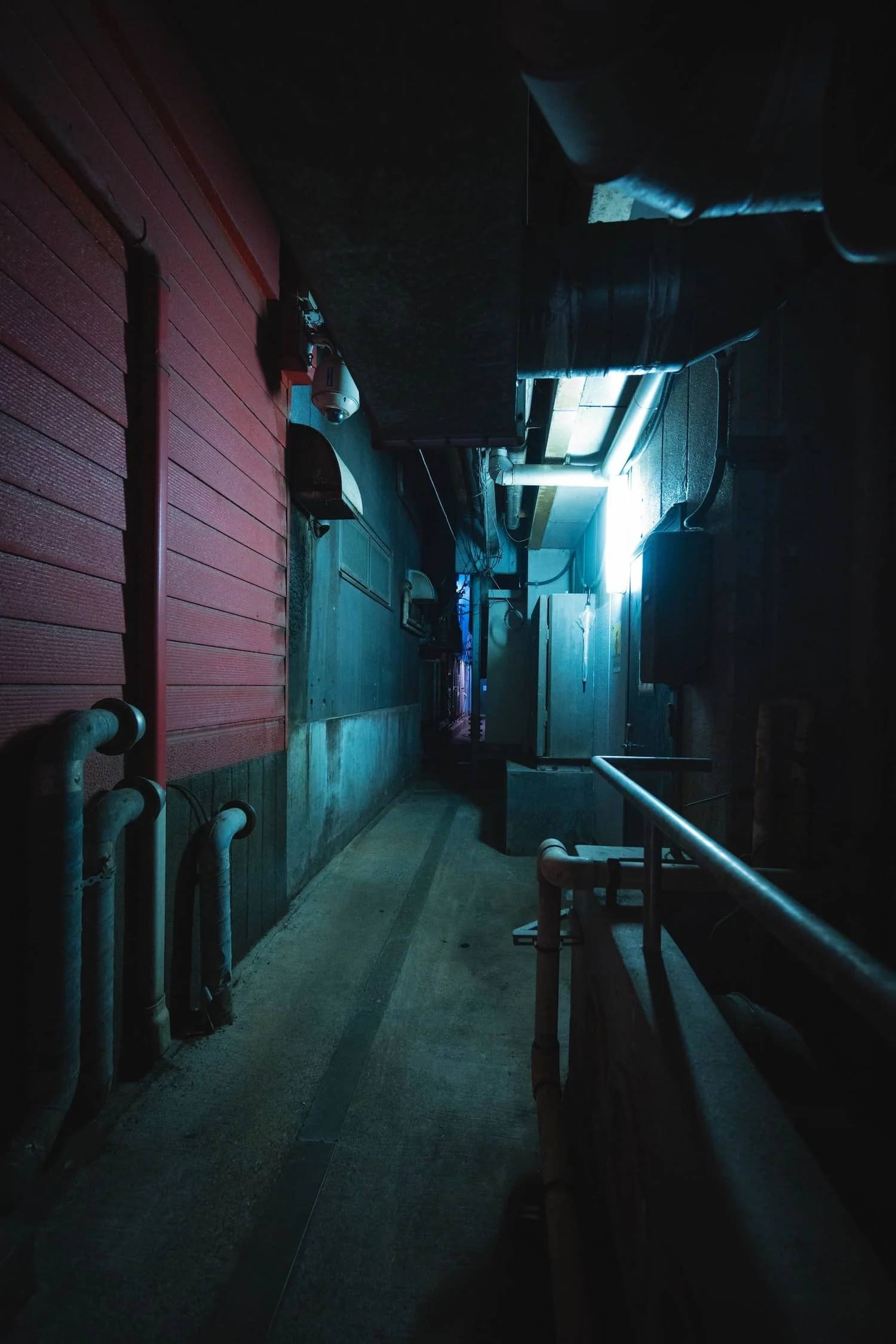 Dark alleyway with pipes and a neon blue light, concrete and wooden walls, and a narrow pathway.