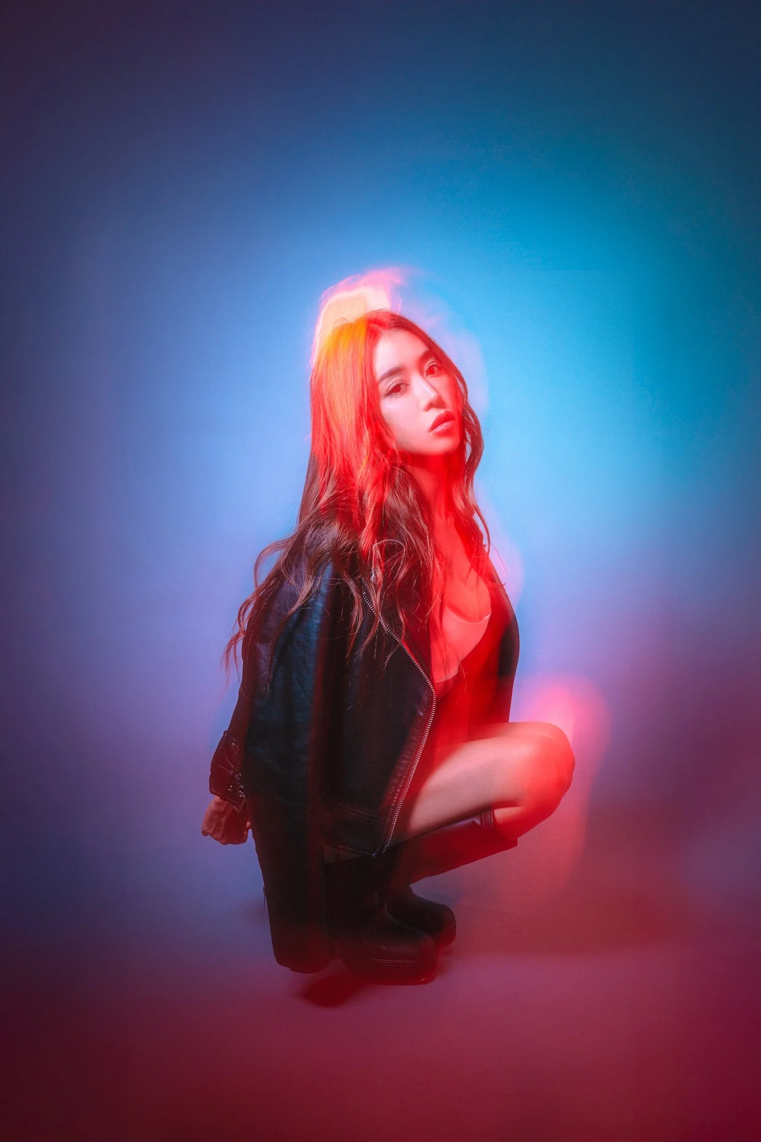 A young woman with long, wavy red hair posing against a gradient background of dark blue and pink hues. She is wearing a black leather jacket and has a serious expression.
