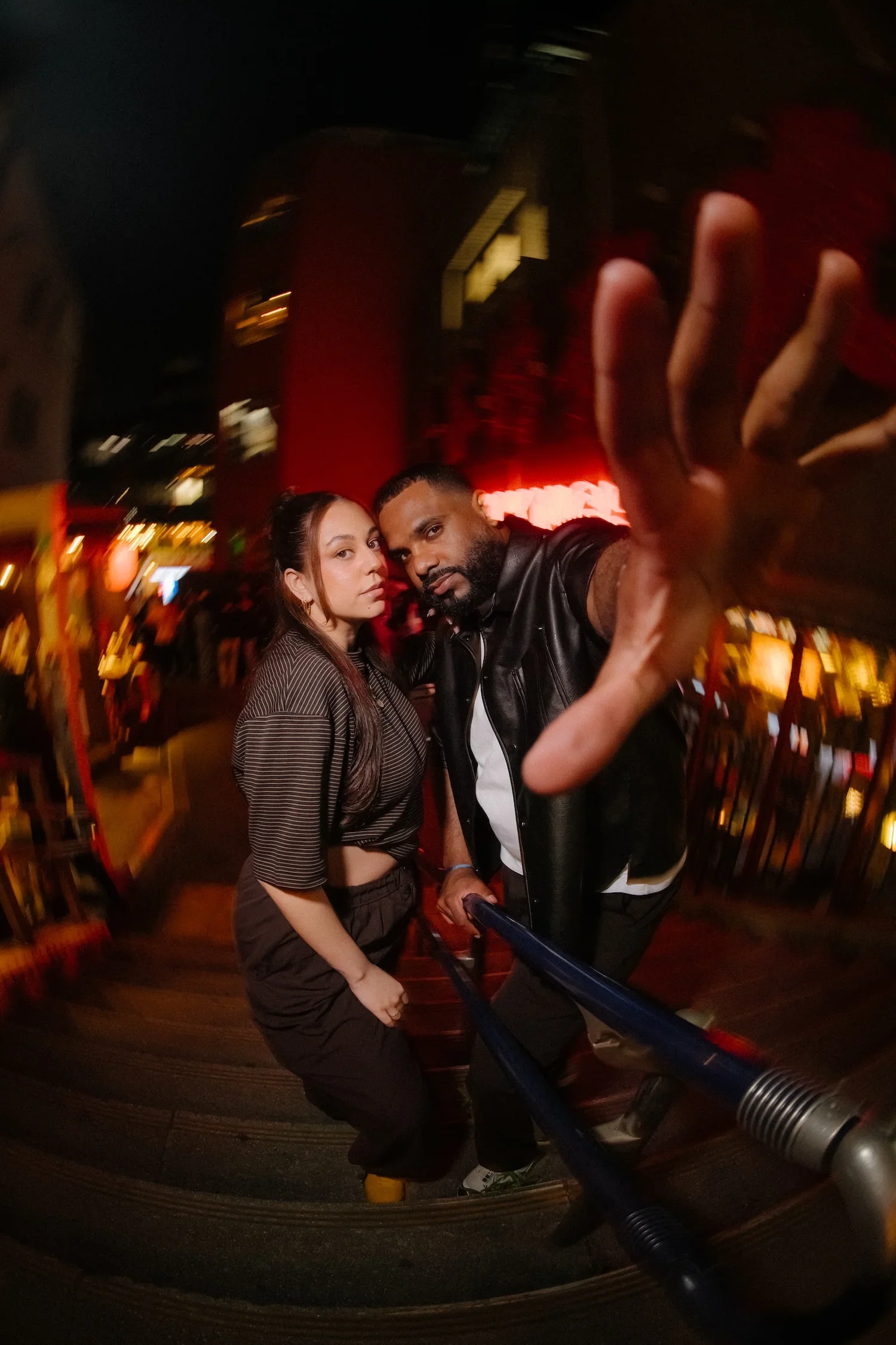 Two people taking a selfie in a dimly lit indoor setting. The woman has long brown hair, and the man has short hair and a beard. They are standing close together with a dark background illuminated by some red and yellow lights.