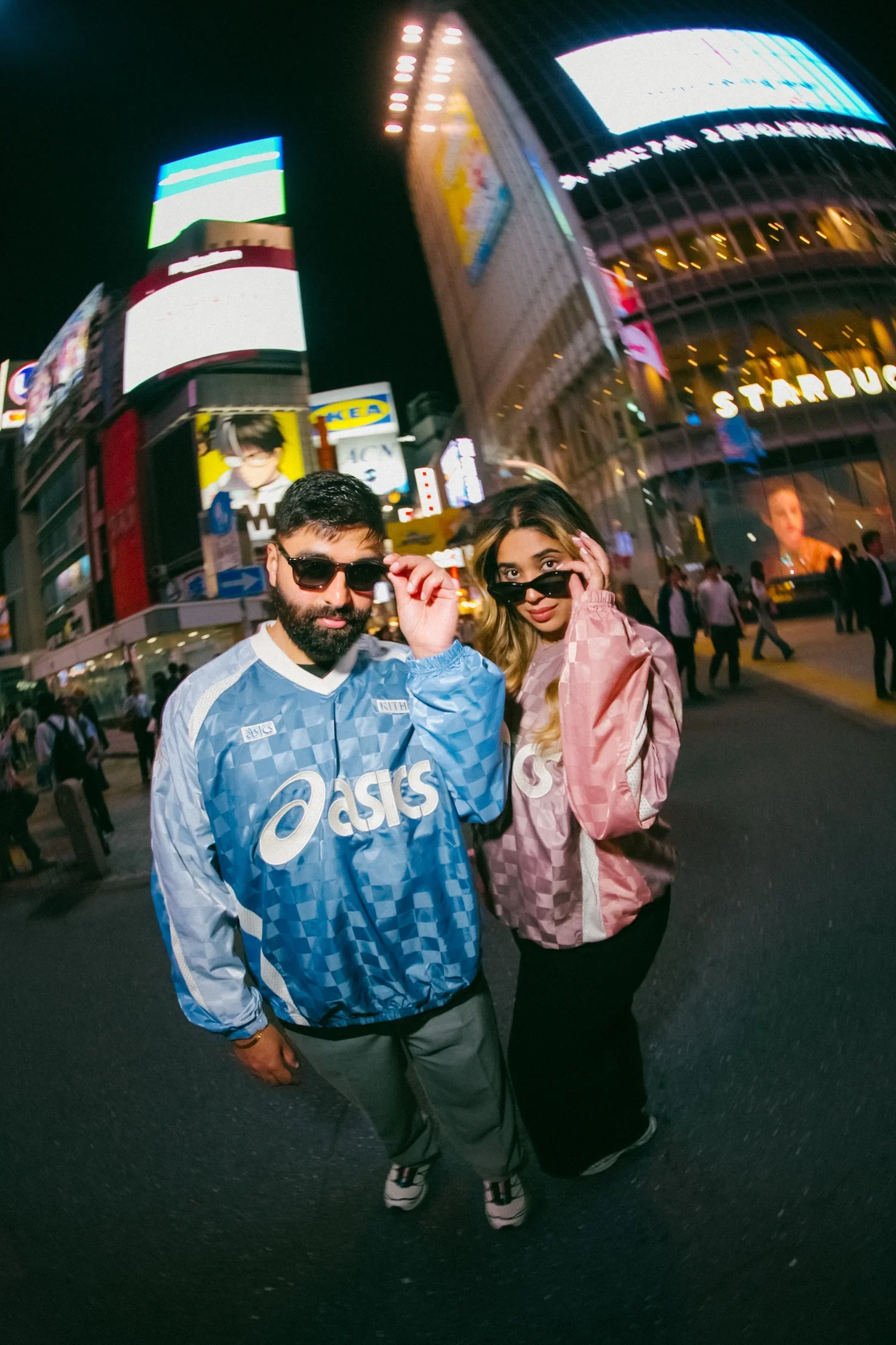 A man and a woman standing in Times Square at night. Both are wearing sunglasses and casual clothing. The background is filled with brightly lit digital billboards and signs, including a Starbucks sign and a billboard with a person’s face on it.