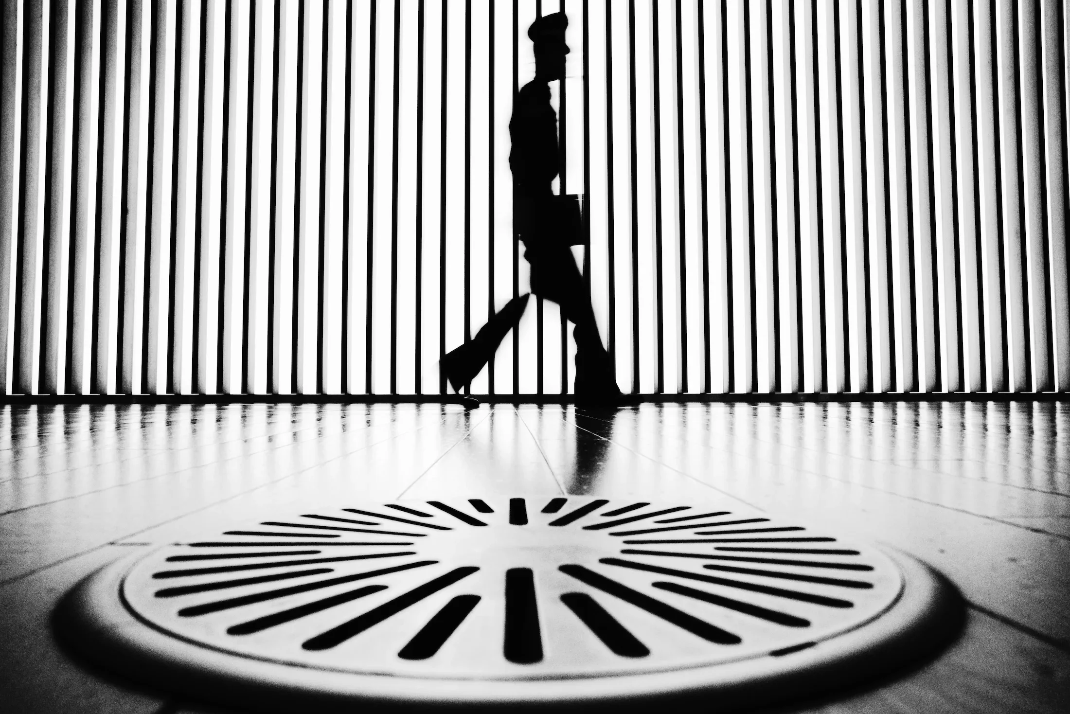 A silhouette of a person walking past a vertical metal fence, with a circular logo on the floor that reads 'VANITY FAIR'.