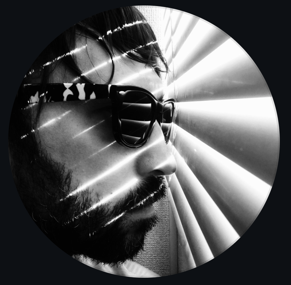 Close-up black and white photo of a man with sunglasses, a beard, and mustache looking through window blinds with light streaming through.