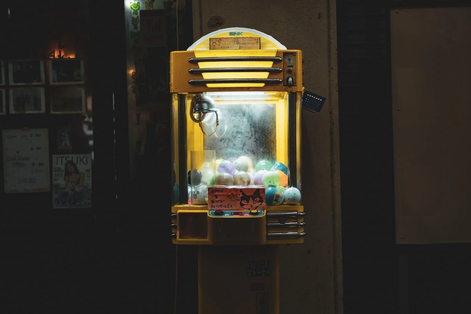 A yellow claw machine with toy balls inside, illuminated in a dark environment, with a pink note featuring Japanese writing and a drawing of a cat's face.