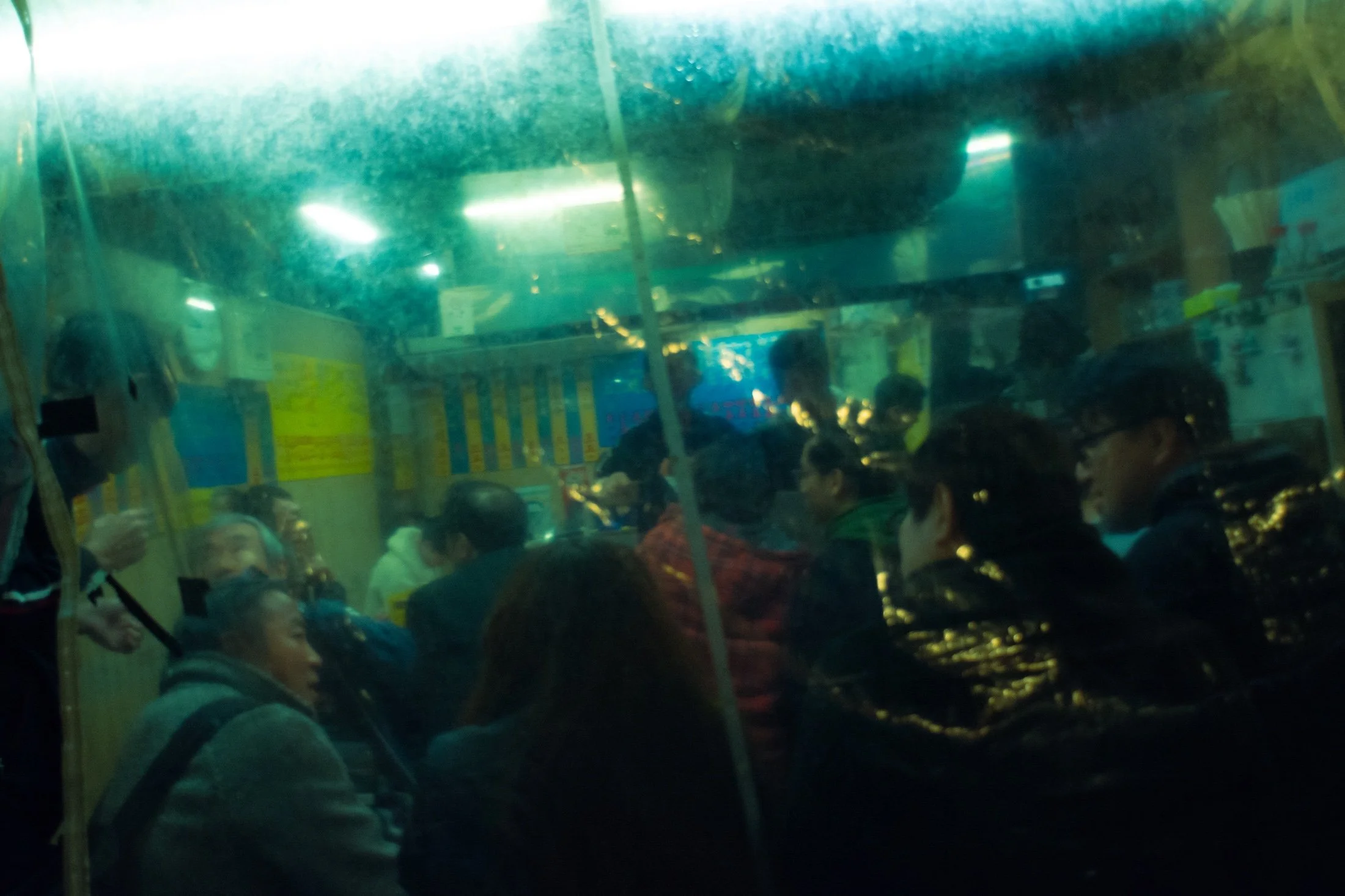 Inside a bus or train with reflections on the window, showing several people seated and a background of shops and signs, indicating a crowded public transport setting.