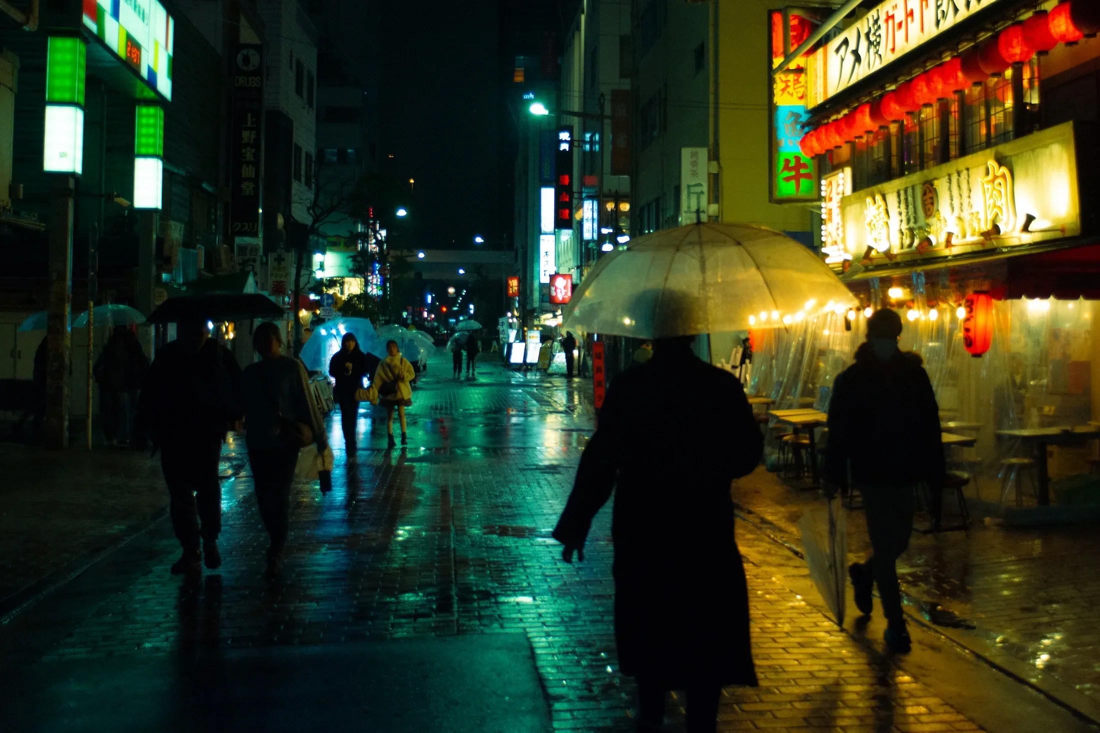 Street scene at night with illuminated signs, people holding umbrellas, and wet pavement reflecting lights in an Asian city.