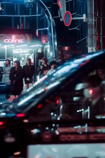 People standing in line outside a modern building with neon lights and a sign that reads 'collins' at night, with cars passing by in the foreground.