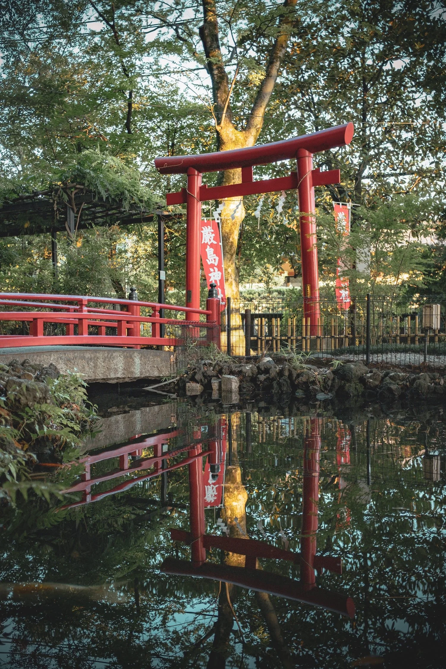 A red Torii gate by a small pond in a green park, with reflections in the water and several banners with Japanese writing.