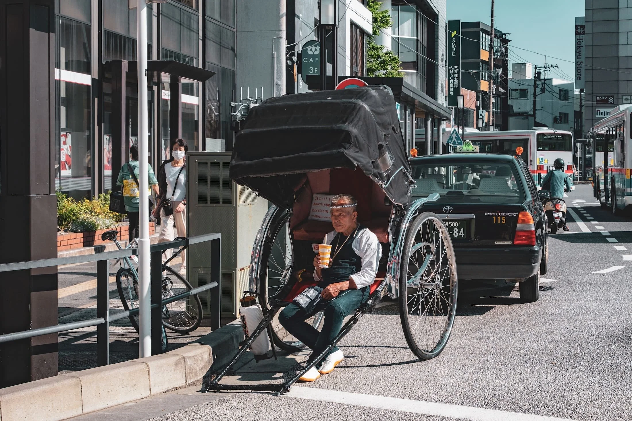 A street scene featuring a traditional rickshaw with an elderly man sitting inside, wearing a face shield and drinking from a cup. People walk past, some wearing masks. In the background, there are buildings, cars, and a bus, indicating an urban environment.