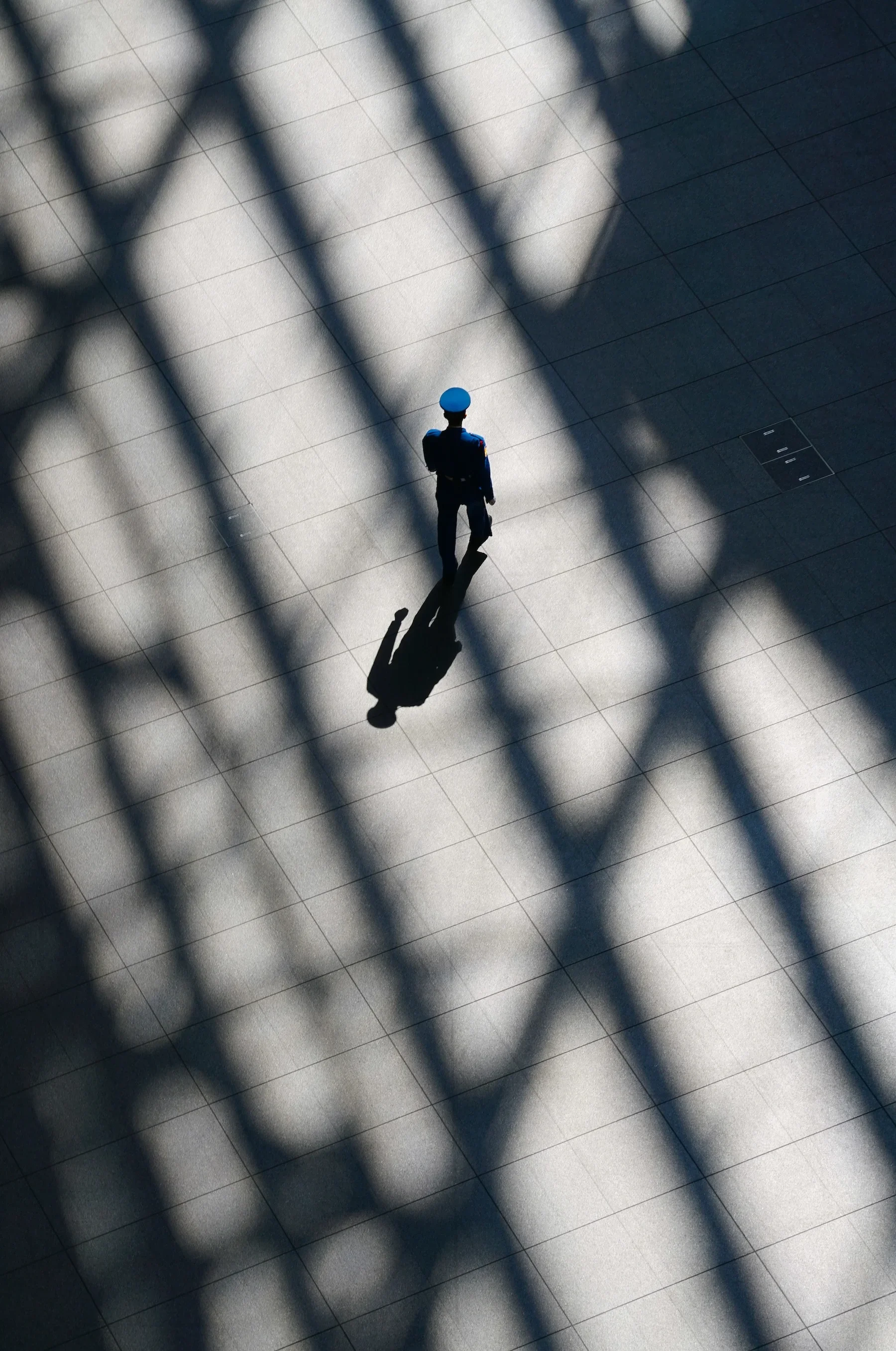 A person in a blue uniform and helmet, possibly a security guard or police officer, walking on a large tiled floor with shadows of a barred structure casting patterns across the surface.