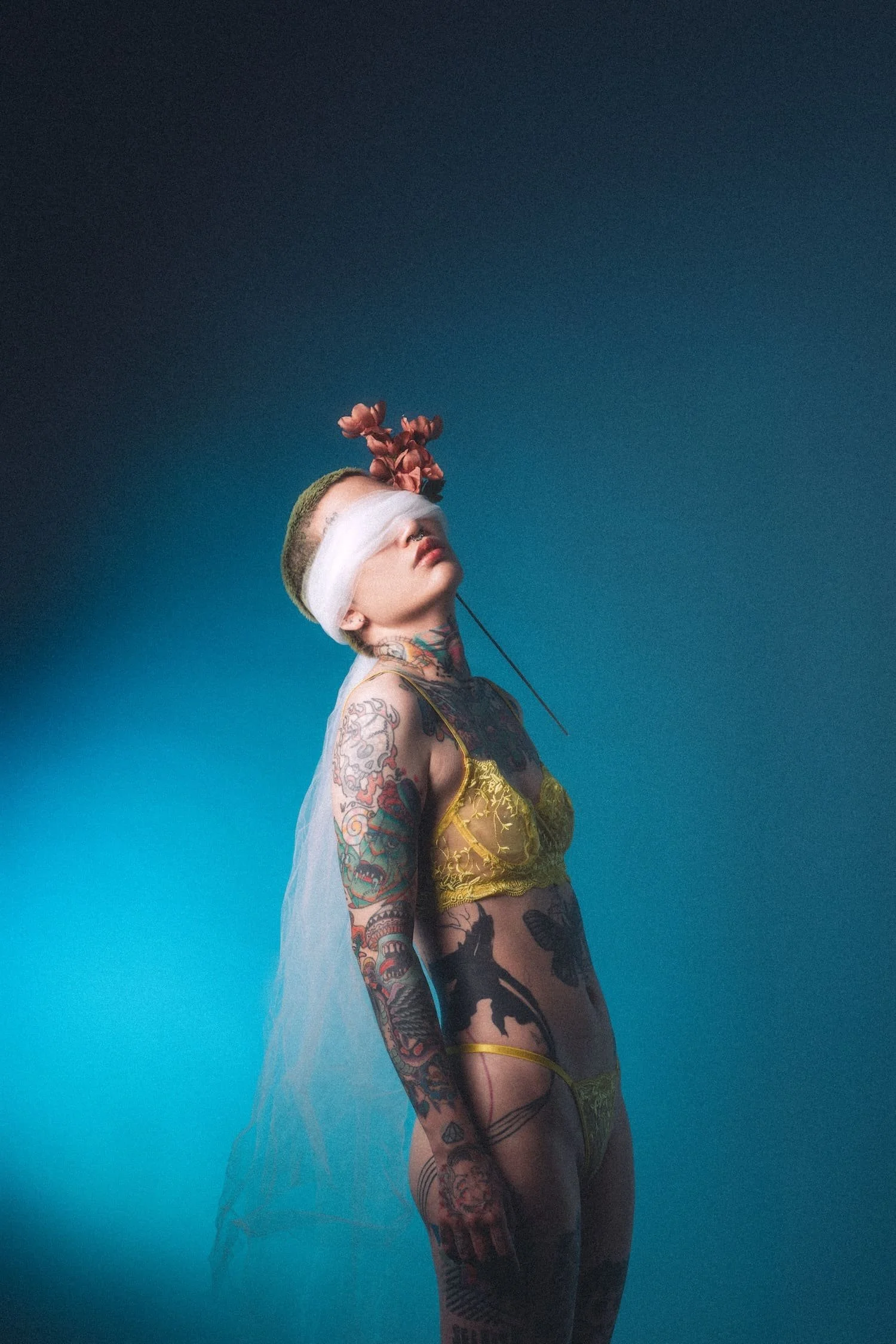 Tattooed woman with a blindfold and a pink flower on her head, wearing yellow lingerie, standing against a blue background.