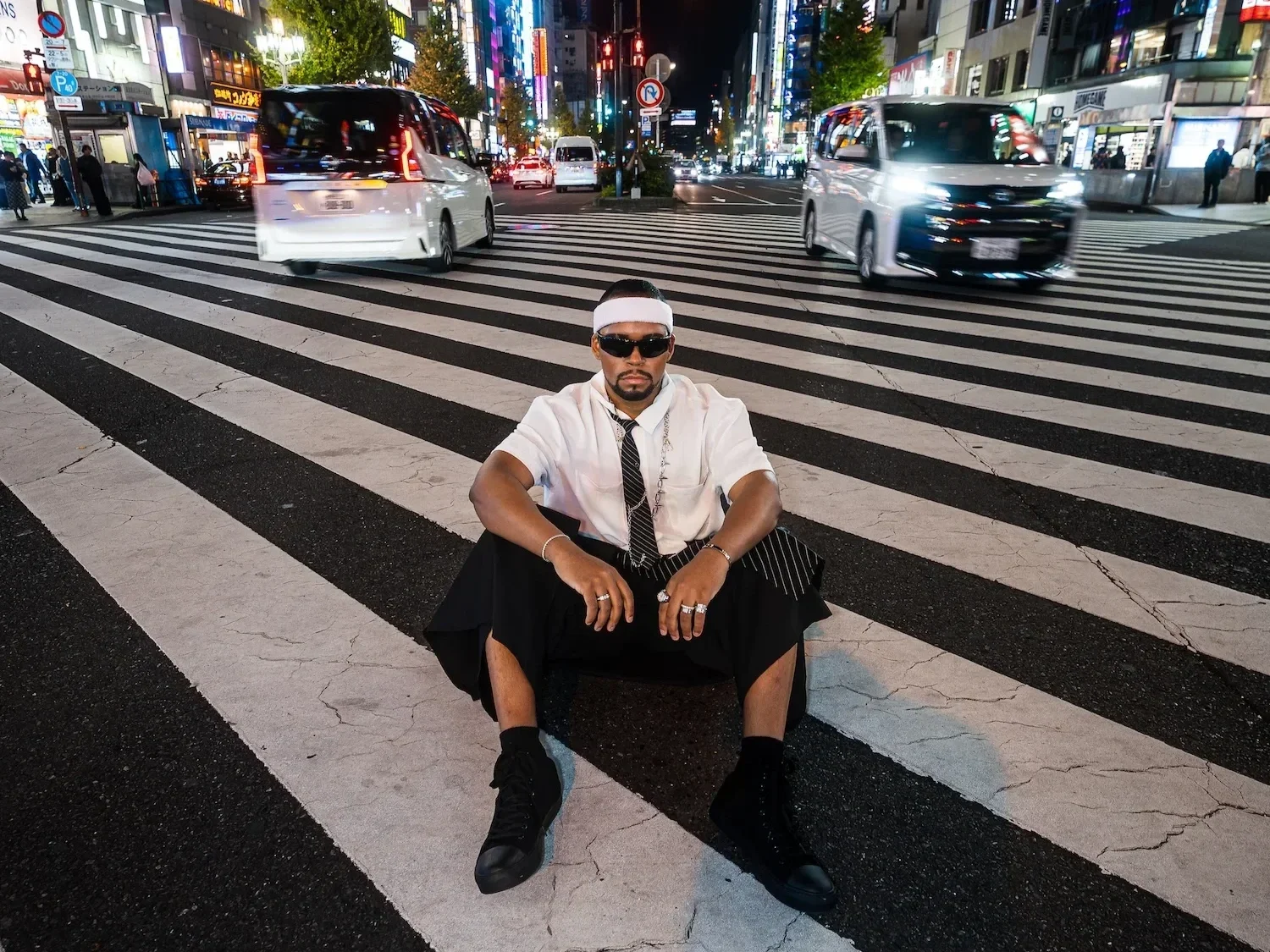 A man dressed in a white shirt, black tie, sunglasses, and black skirt sits on a crosswalk in an urban area at night, with city lights, cars, and buildings in the background.