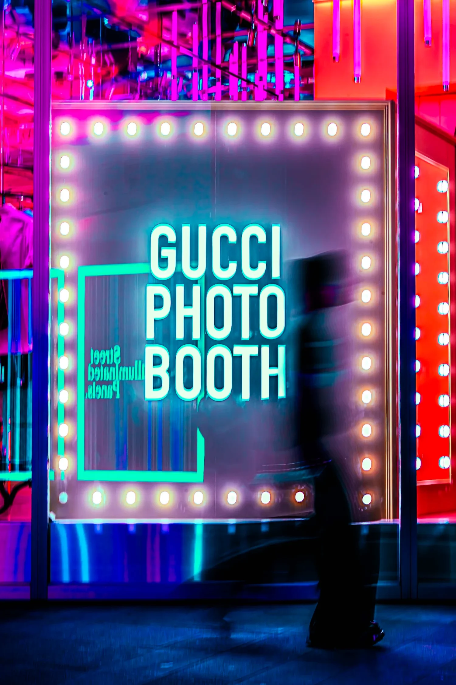A person standing in front of a brightly lit mirror photo booth with neon lights, saying 'GUCCI PHOTO BOOTH' in a vibrant nightlife setting.