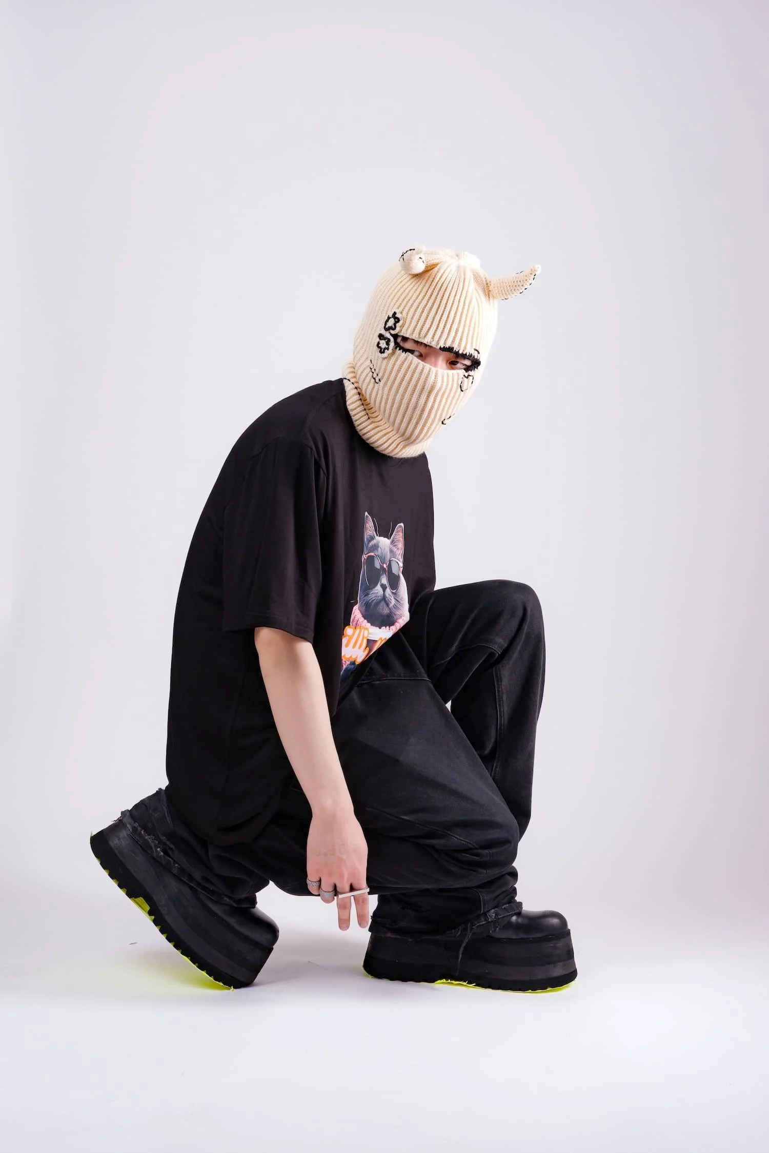 Studio fashion portrait of a model crouching in chunky platform shoes while wearing a beige horned knit mask and oversized graphic t-shirt, photographed against a clean white backdrop by Tokyo photographer Deniz Demir.