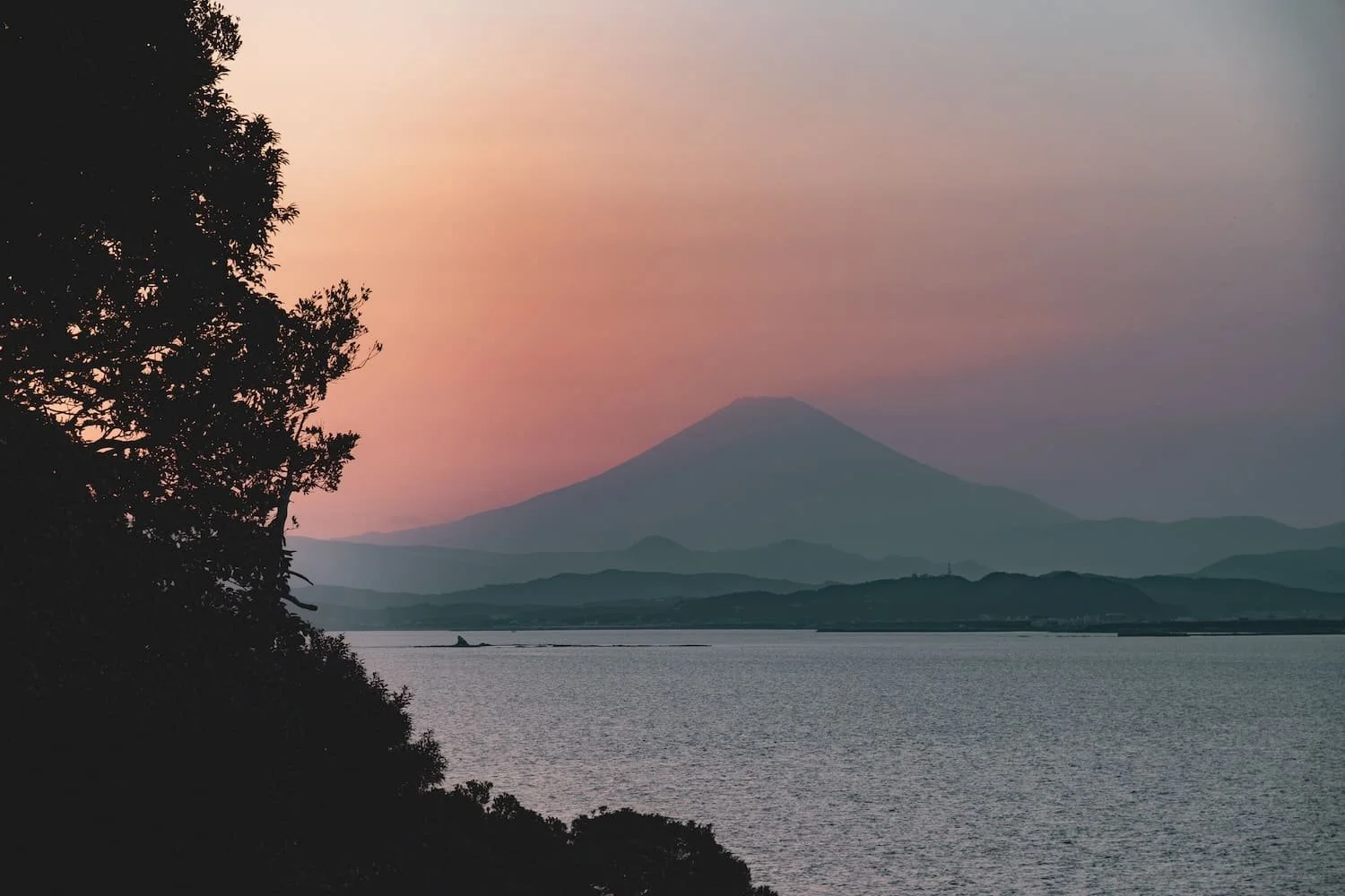 Scenic view of a mountain, likely Mount Fuji, at sunset or dawn with soft pink and purple sky, framed by dark trees and calm water in the foreground.