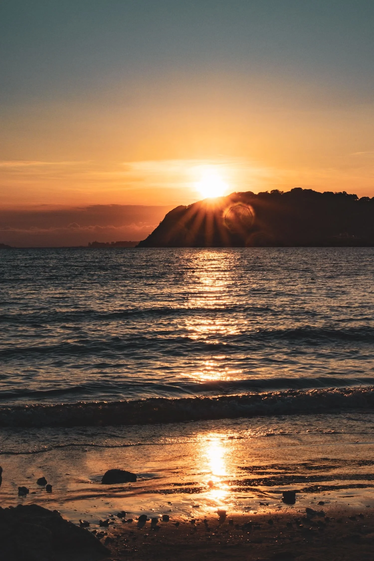 Sunset over the ocean with a silhouetted hill, reflecting golden light on the water and wet sand.
