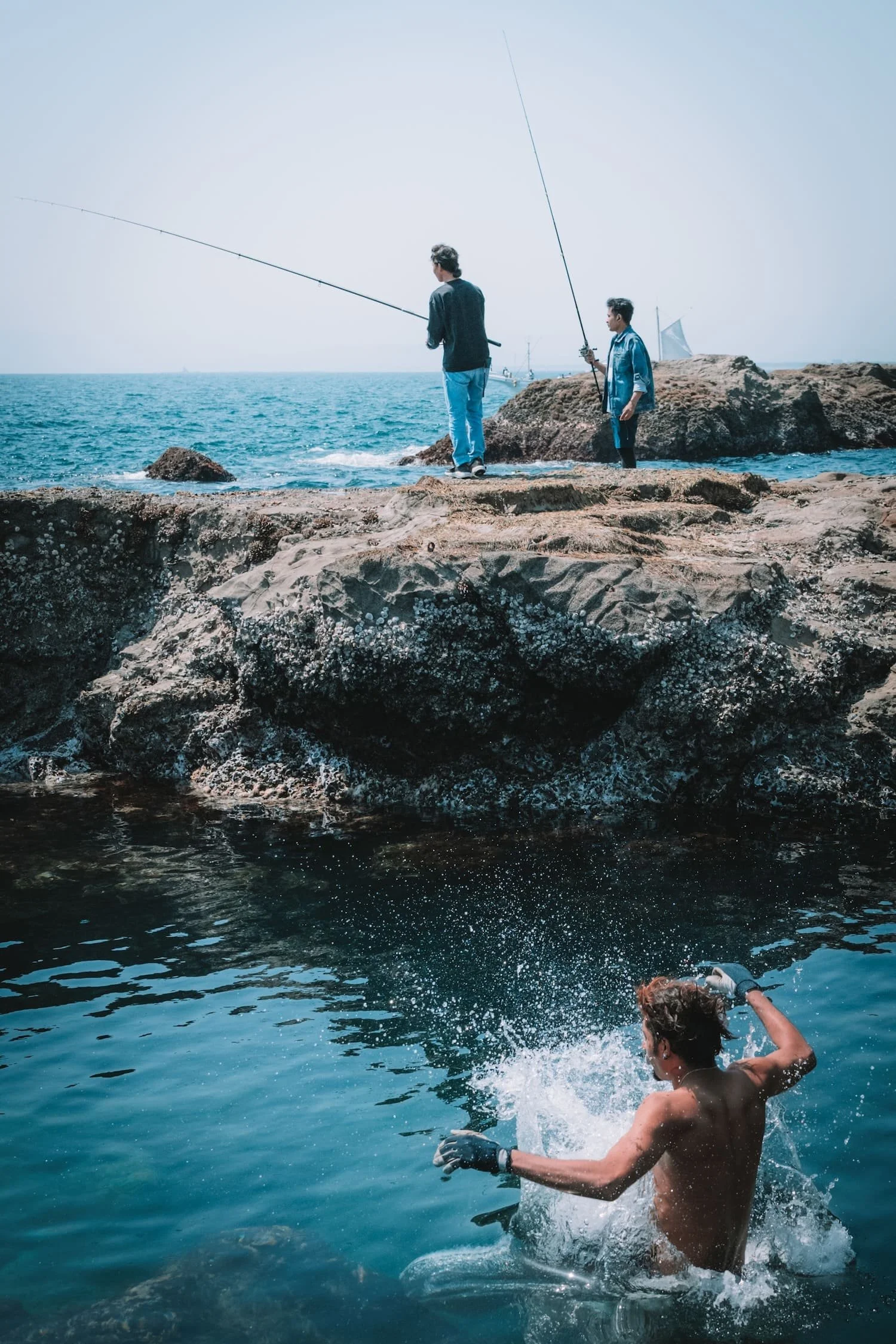 Two men fishing on rocks by the ocean, with one person swimming in the water below.