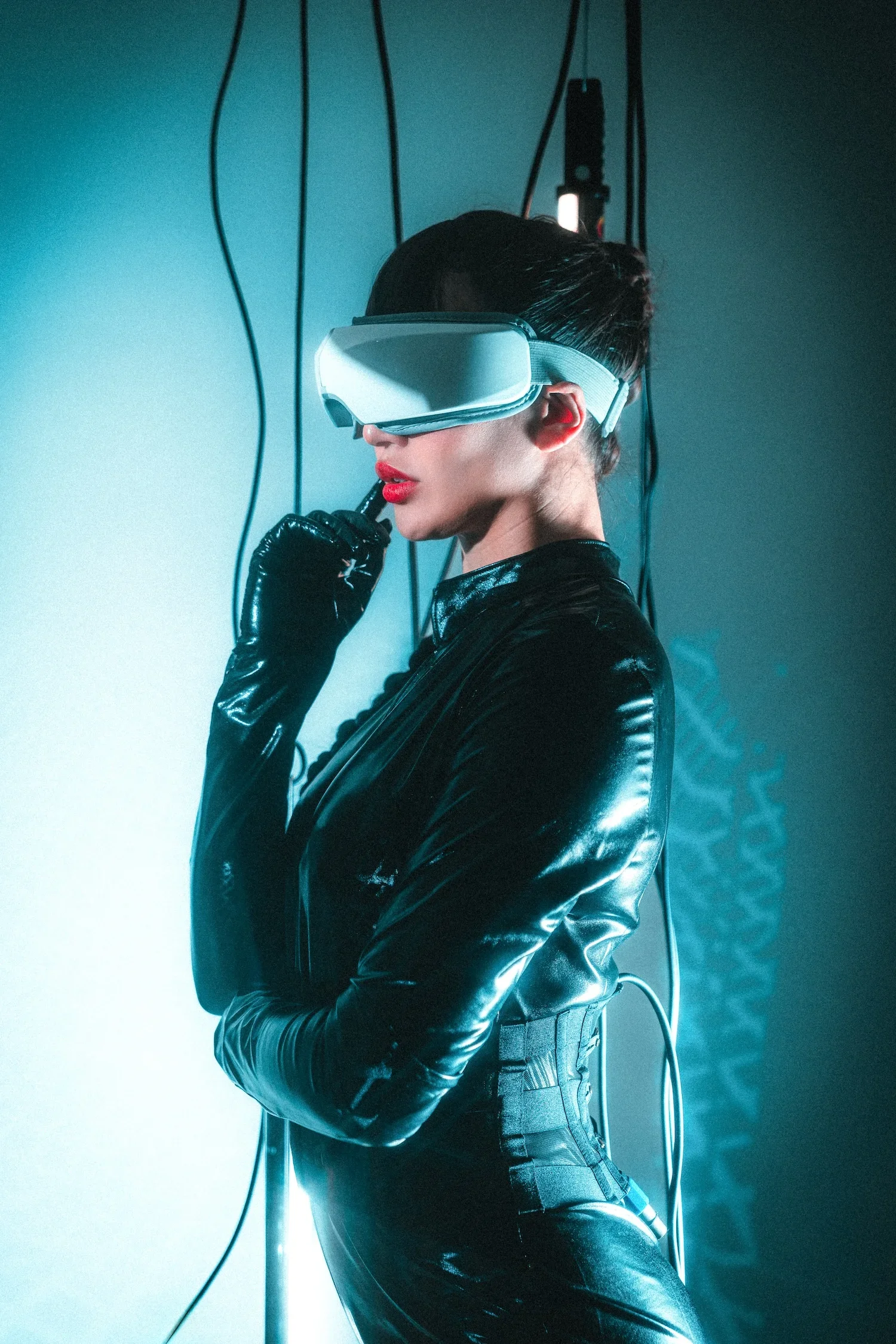 A woman wearing a virtual reality headset, glossy black leather outfit, and red lipstick, standing next to a wall with black cables hanging, with blue lighting creating a futuristic atmosphere.
