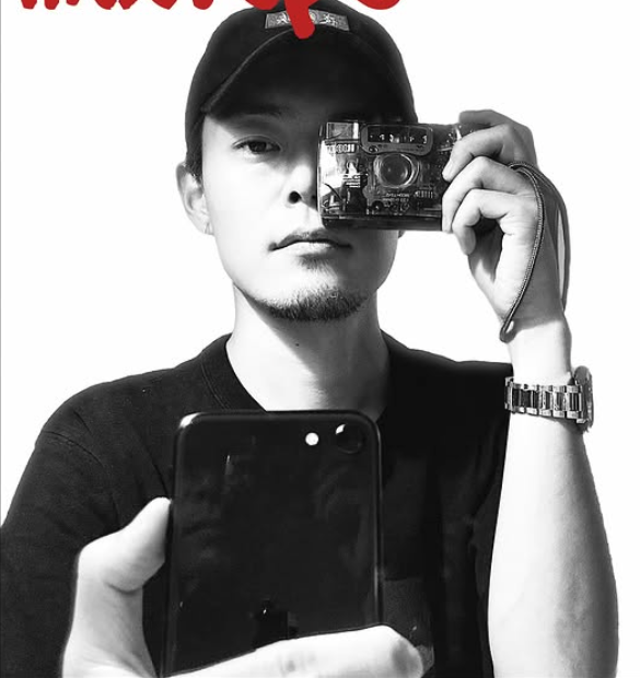 Young man taking a mirror selfie with a smartphone, wearing a black cap and a watch, with another person holding a camera in front of him.