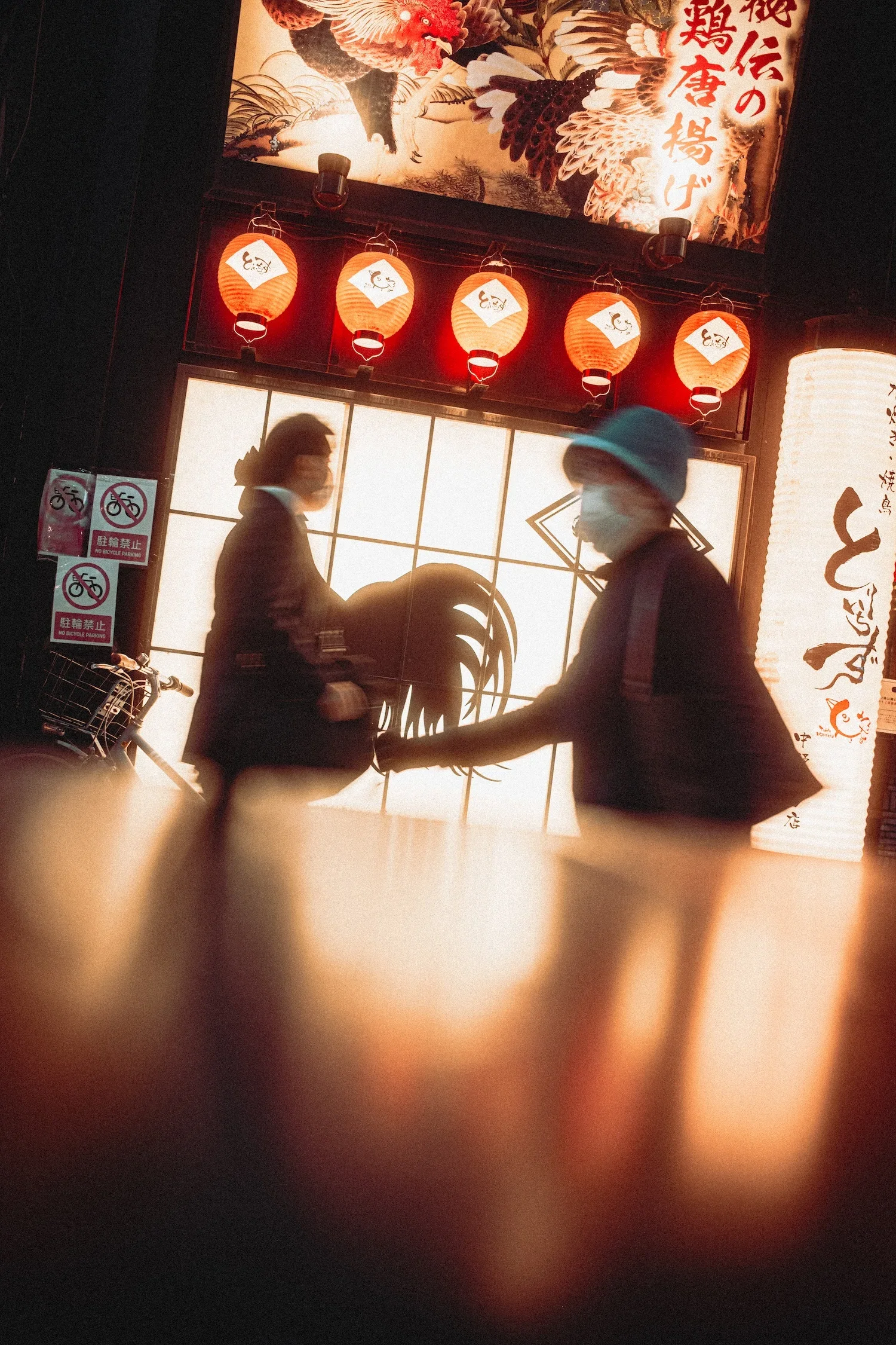 Two people are walking past a Japanese restaurant illuminated by red lanterns and a large illuminated sign featuring traditional Japanese art and calligraphy. The image is slightly blurry with a finger partially covering the camera lens in the foreground.
