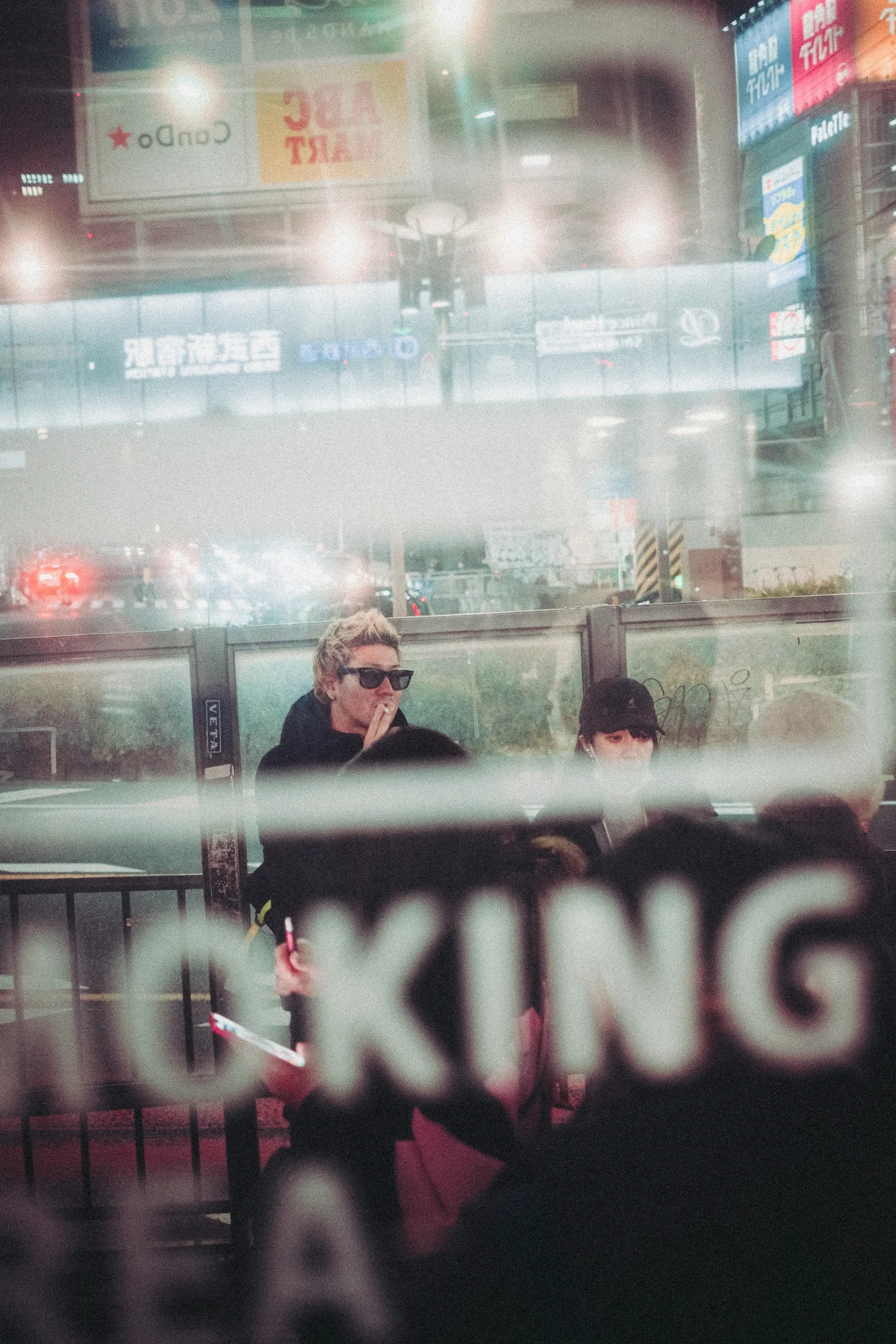 Nighttime city scene seen through a glass window with reflections of bright advertisements and lights, and two people sitting at a bus stop or outdoor seating area.