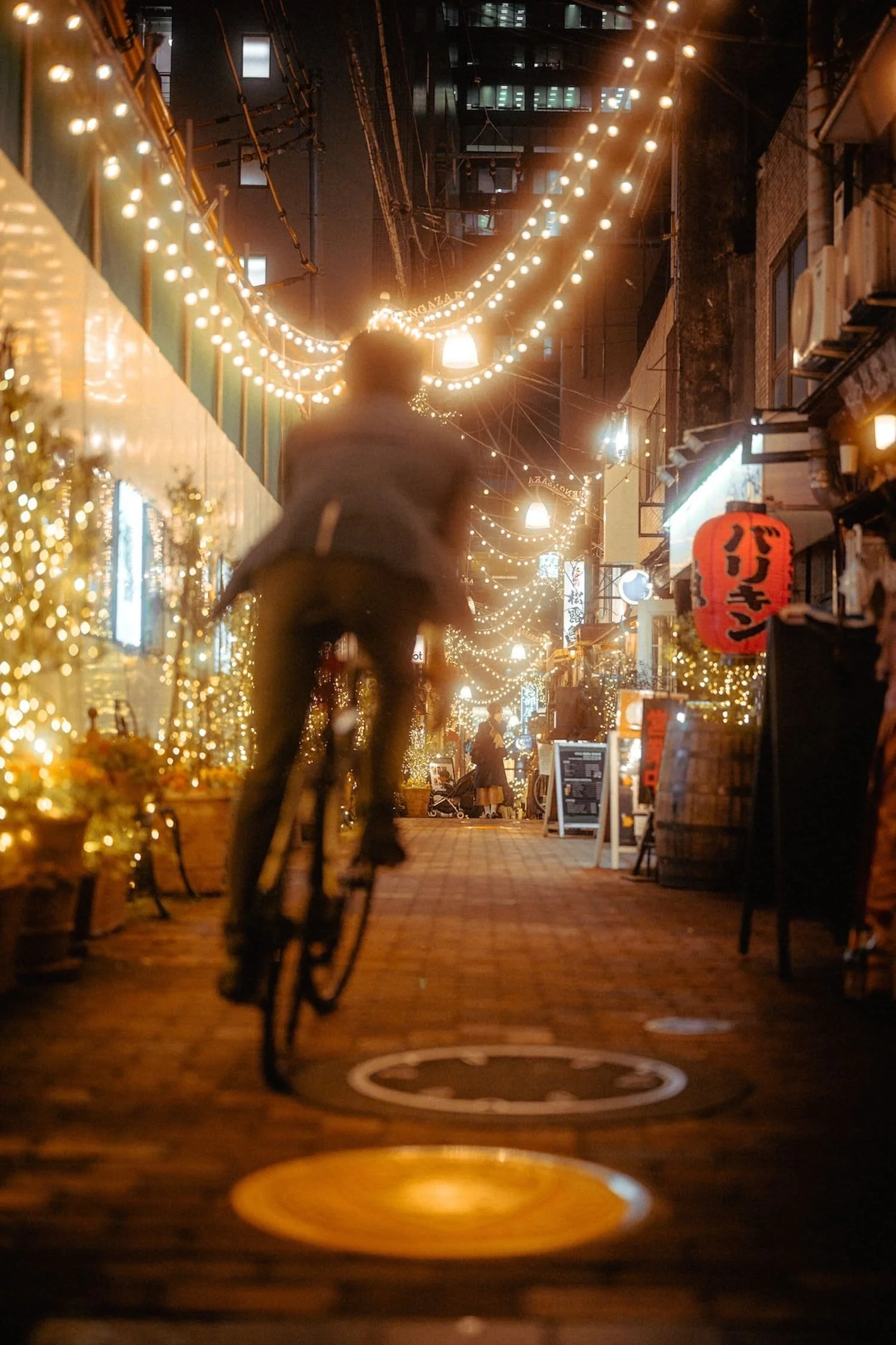 A person riding a bicycle down a warmly lit urban alley at night, decorated with string lights and festive signs.