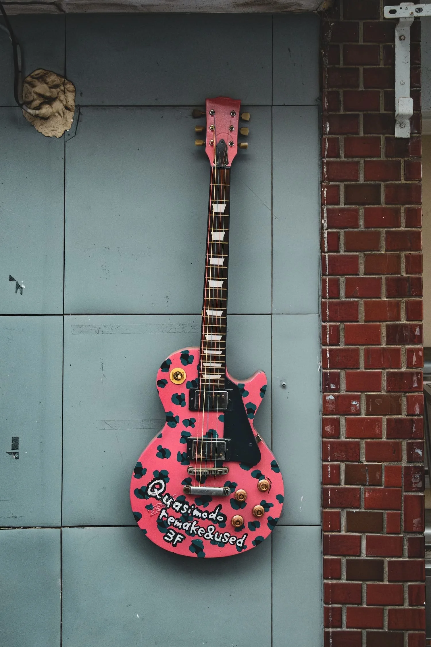 Pink electric guitar with black and blue spots, mounted on a gray wall next to a red brick wall. The guitar has a handwritten message: "@quasmado.remaked&used 3F".