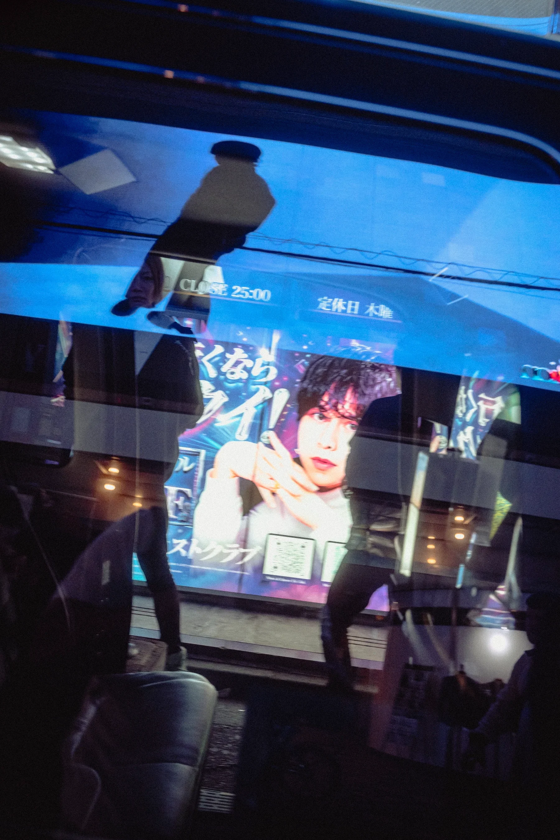 A reflection of a woman on a glass storefront, with a large advertisement for a male model displaying text in Japanese and English, showing a city street scene.