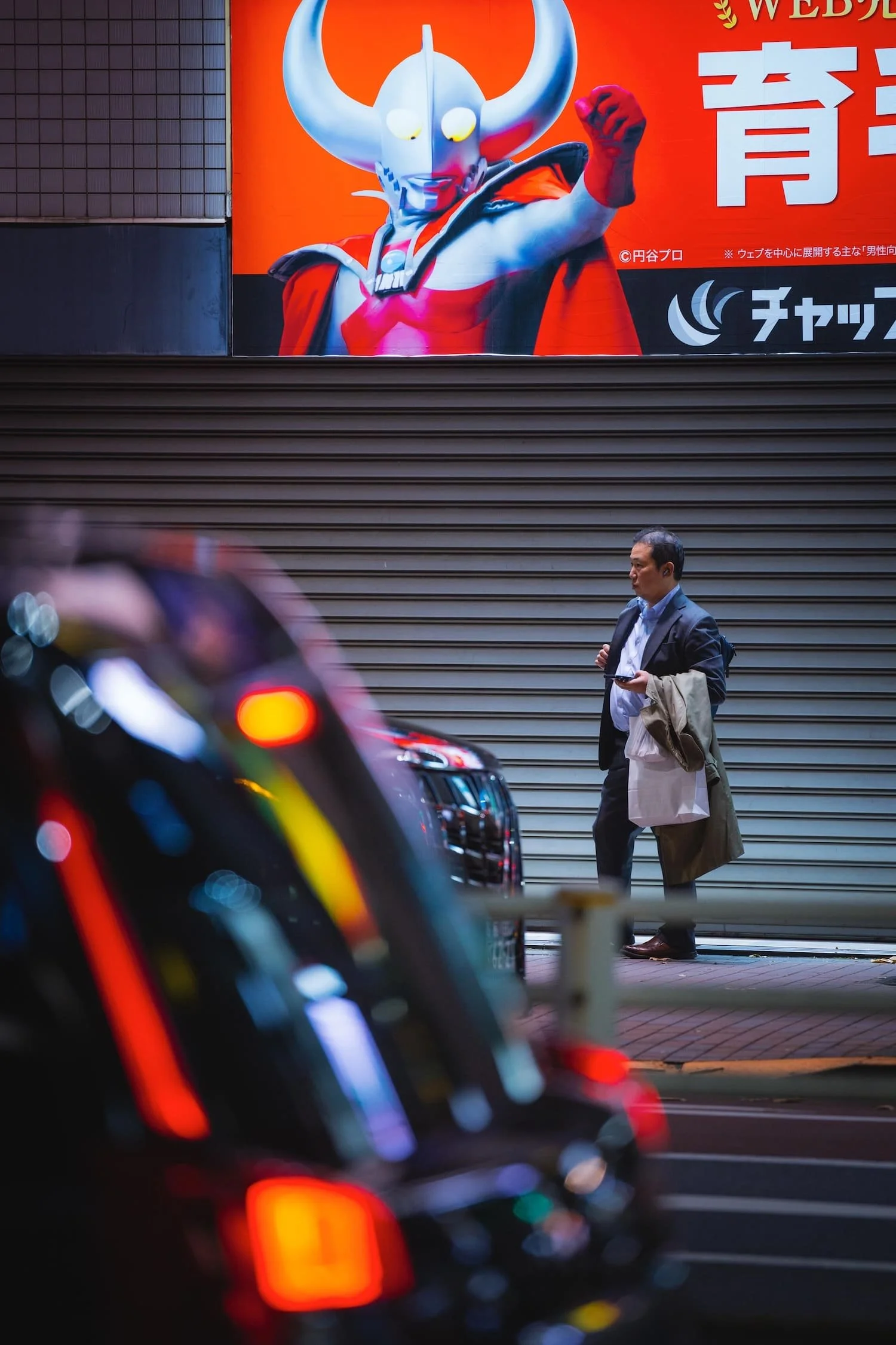 A man in a suit walking past a closed store with a large billboard of a superhero character with horns, wearing a red and black costume, and making a fist.