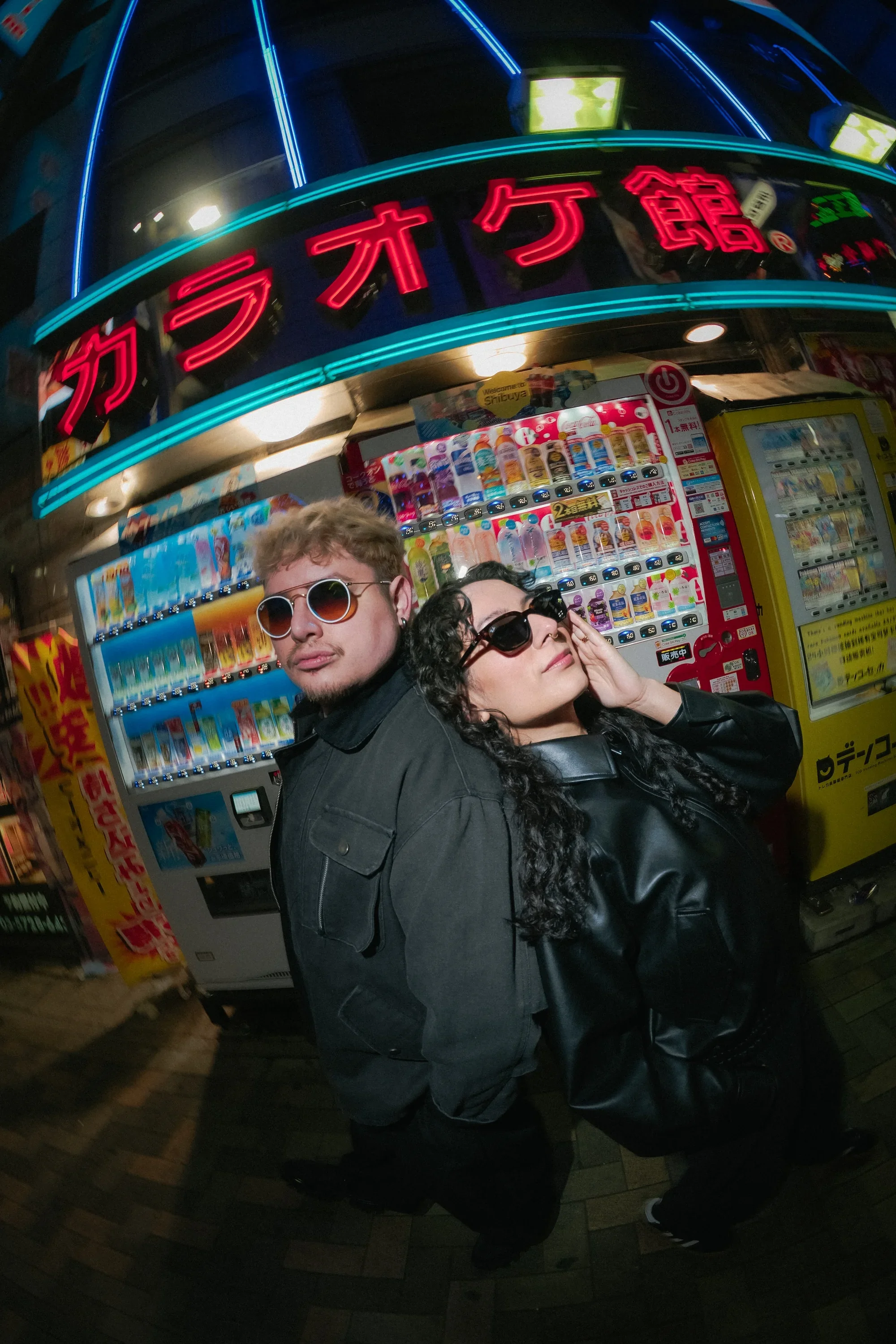 Two people posing in front of a vending machine at night in an outdoor urban setting. They are wearing dark clothing and sunglasses, with a neon-lit building sign overhead with Japanese characters.