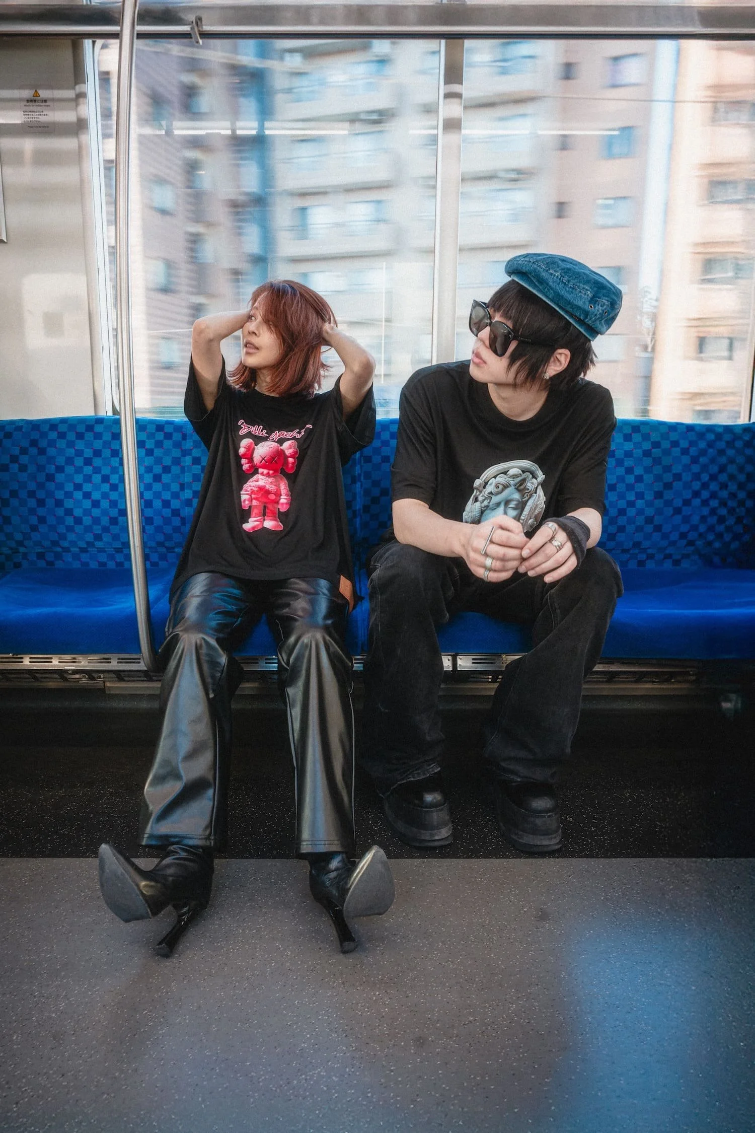 Two models sitting across from each other inside a Tokyo subway carriage wearing oversized graphic streetwear t-shirts, captured through a moving train window with city buildings blurred outside, photographed by Tokyo photographer Deniz Demir. Shot f