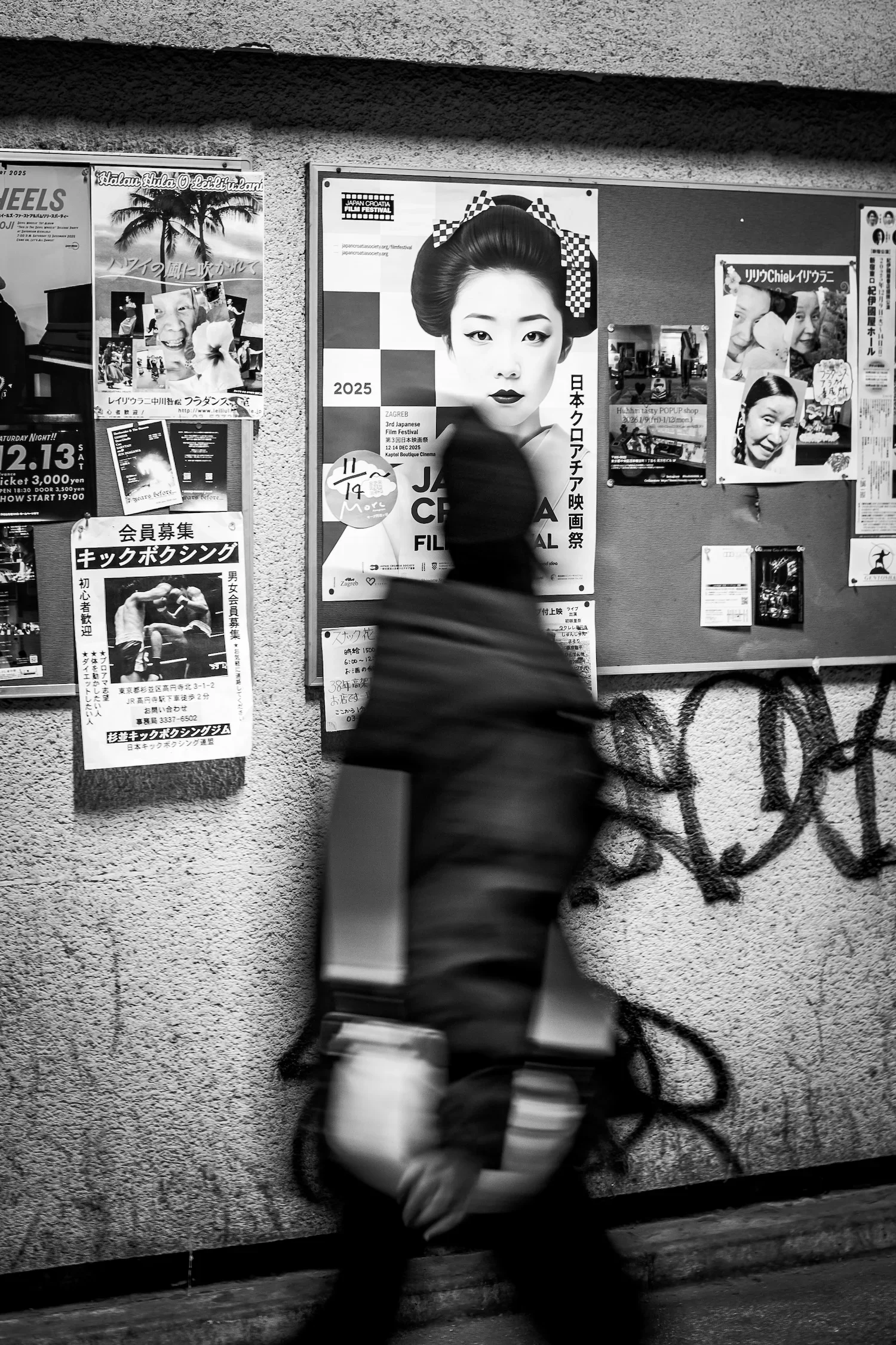 A blurred person walking past a bulletin board with various posters and flyers on a textured wall.