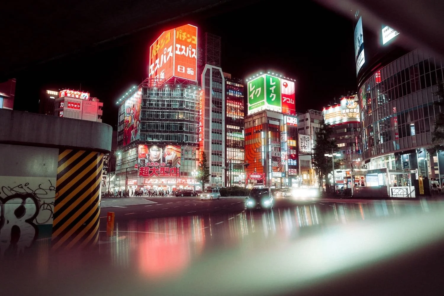 Night view of a brightly lit city street with tall buildings covered in colorful neon signs, reflections on wet pavement, and some cars driving by.