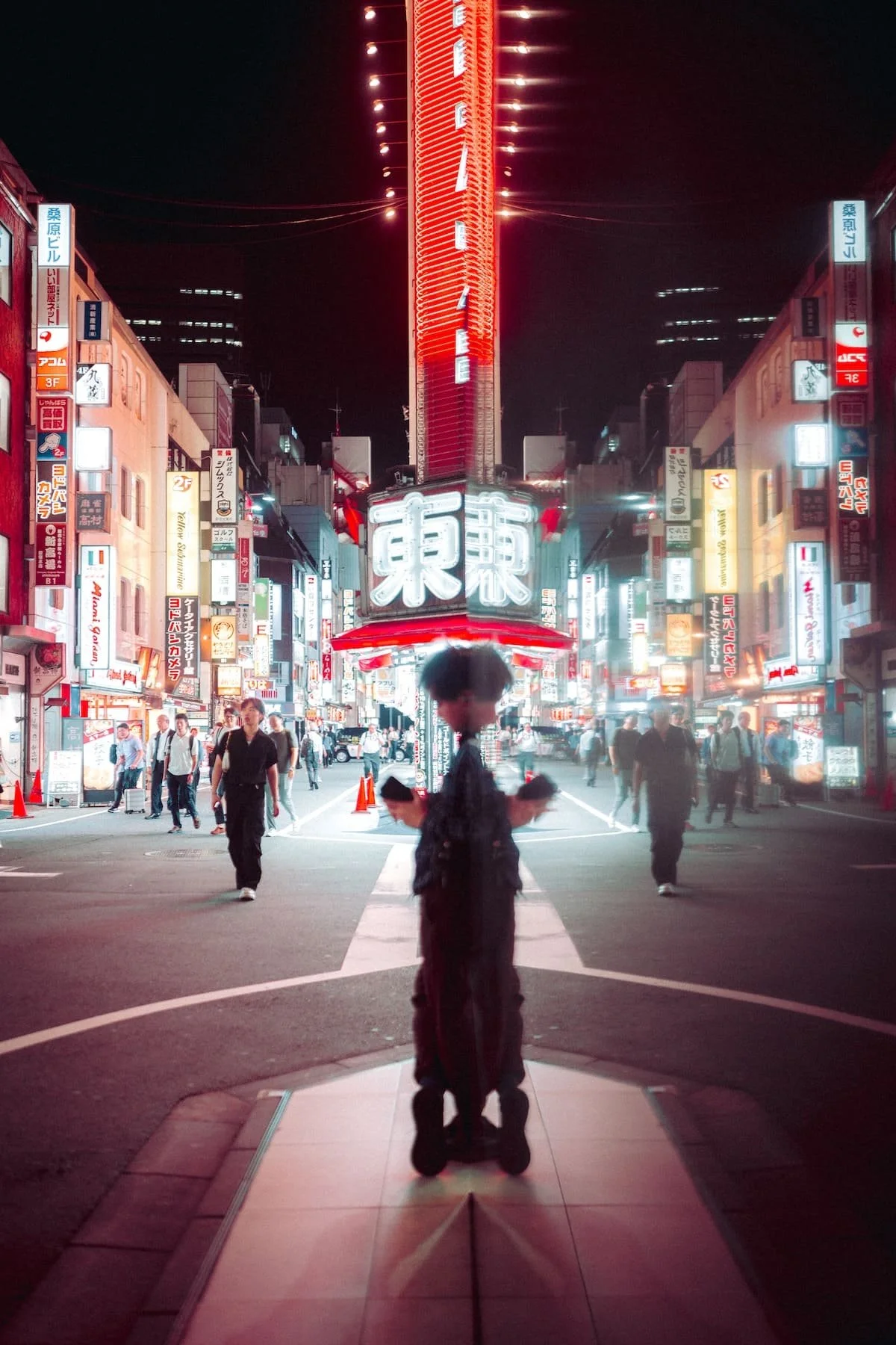 Nighttime city street scene in Tokyo with bright neon signs, many pedestrians, and a person standing in the foreground using a smartphone.