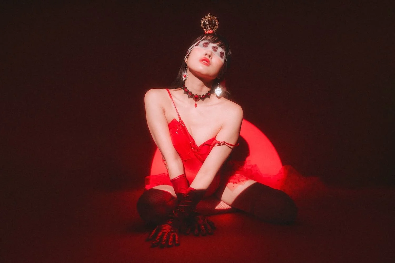A woman in a red dress and black thigh-high stockings kneeling in front of a glowing red circle, wearing jewelry and a crown, with dramatic makeup and lighting.