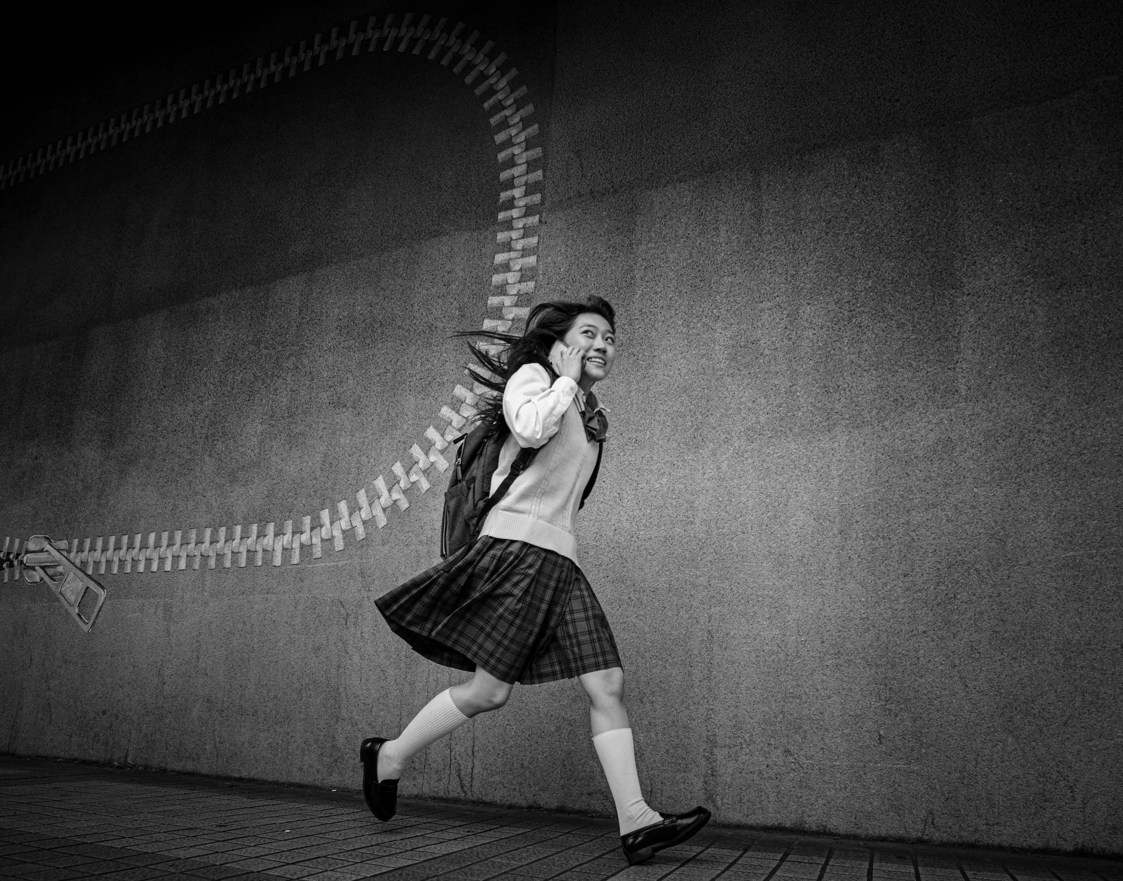 A young woman with long hair is running and holding a phone to her ear, smiling. She is dressed in a school uniform with a plaid skirt, white knee-high socks, and black shoes, and carries a backpack. She is running against a textured wall with a curved brick pattern, and the photo is in black and white.