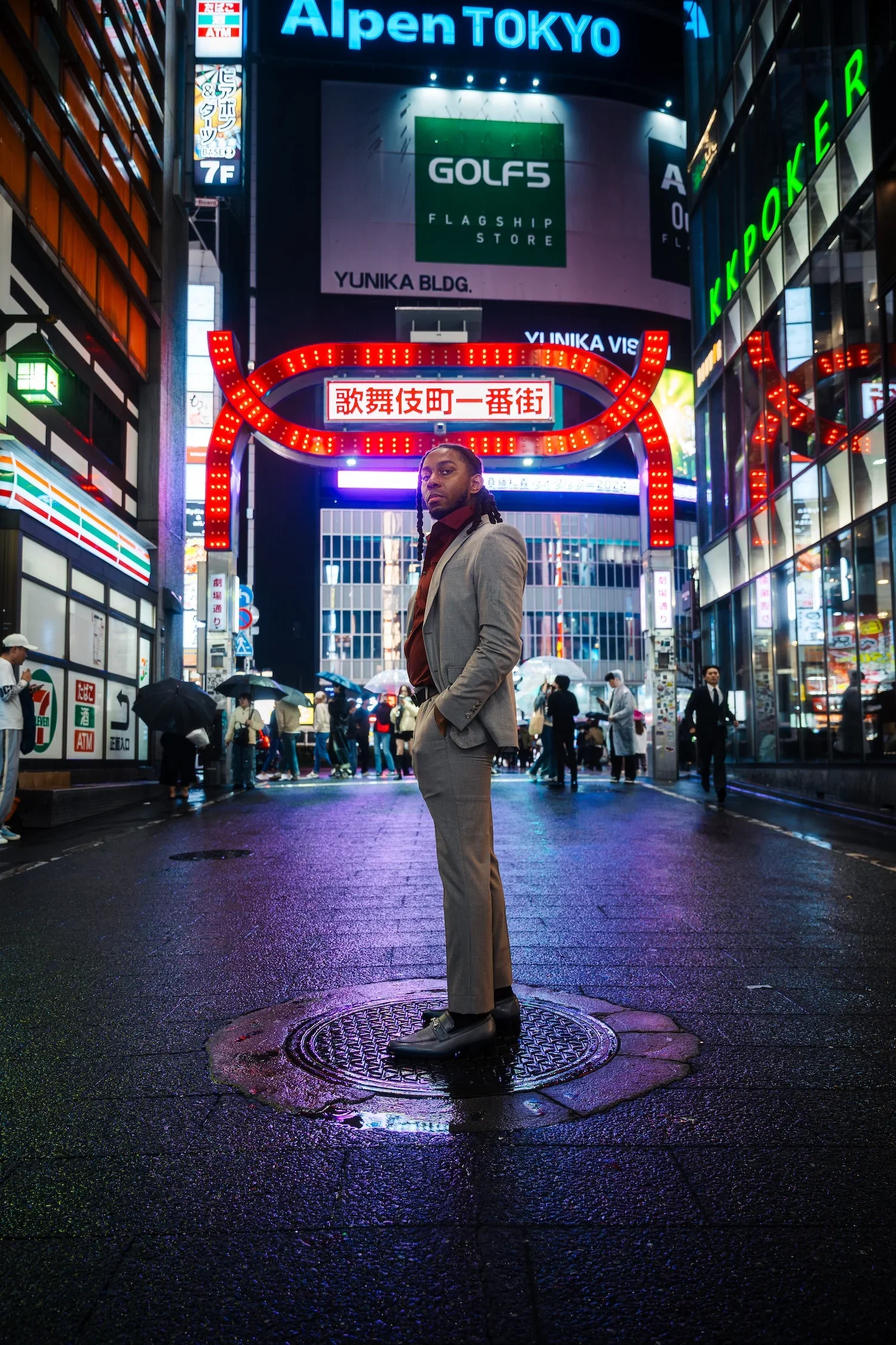 A man with braided hair wearing a beige suit and dark shirt standing on a wet city street at night, illuminated by colorful neon signs and advertisements in Tokyo, Japan.
