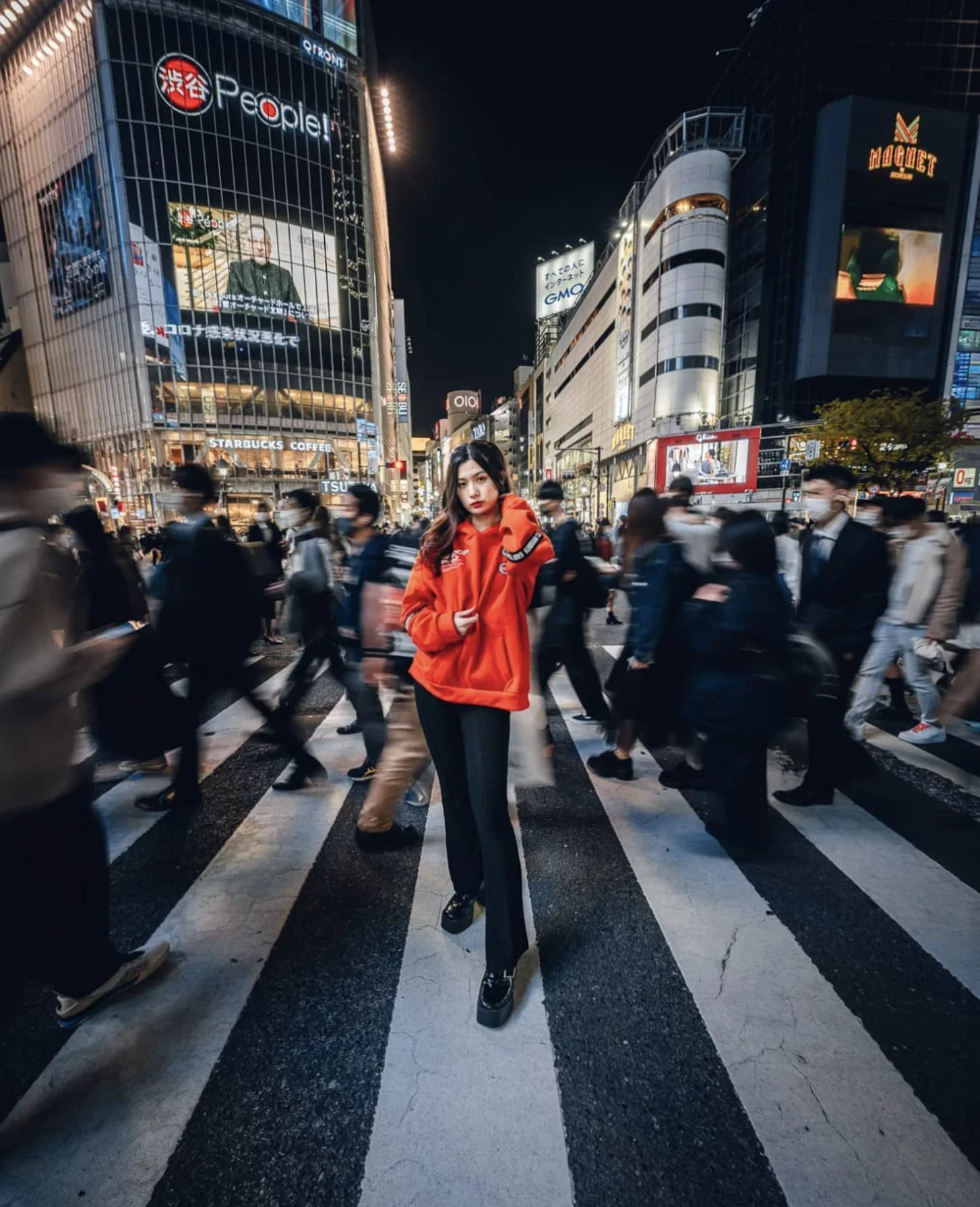 A woman in a red hoodie and black pants standing in the middle of a busy city street at night with illuminated buildings and billboards, surrounded by blurred pedestrians.