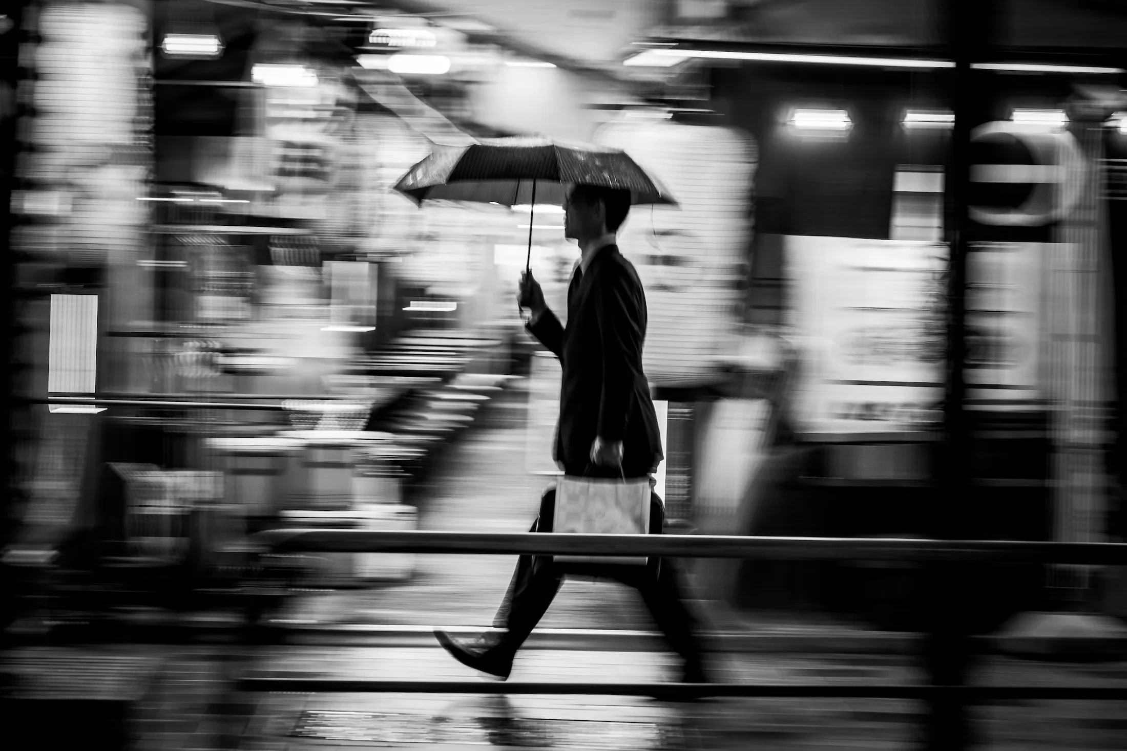 A man in a suit walking with a shopping bag while holding an umbrella in a blurred city street scene at night.