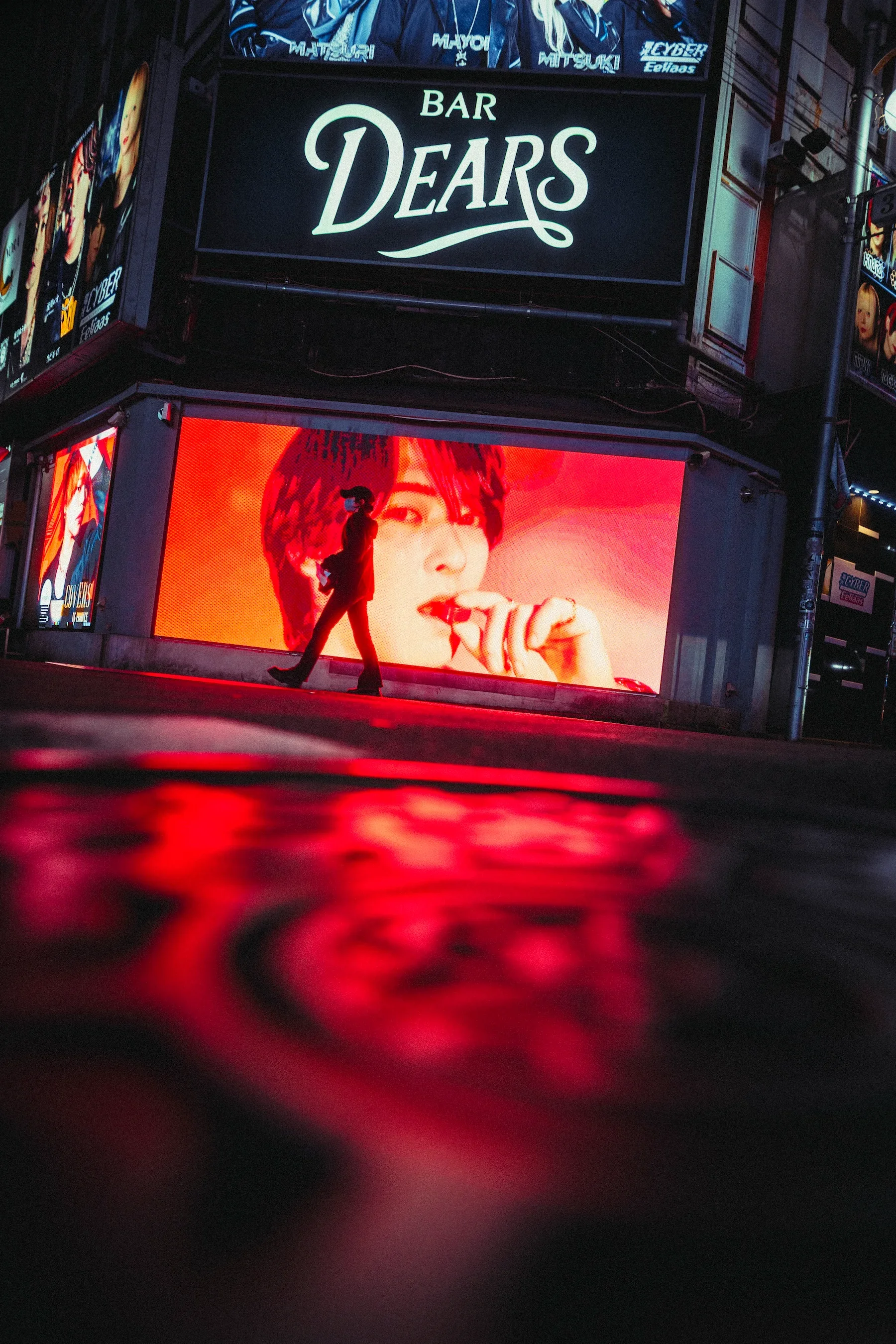 The image shows a person walking in front of a large digital advertisement screen with a close-up of a man's face. Above the screen, there are signs for a bar named 'Dears' and other advertisements in an urban setting at night.