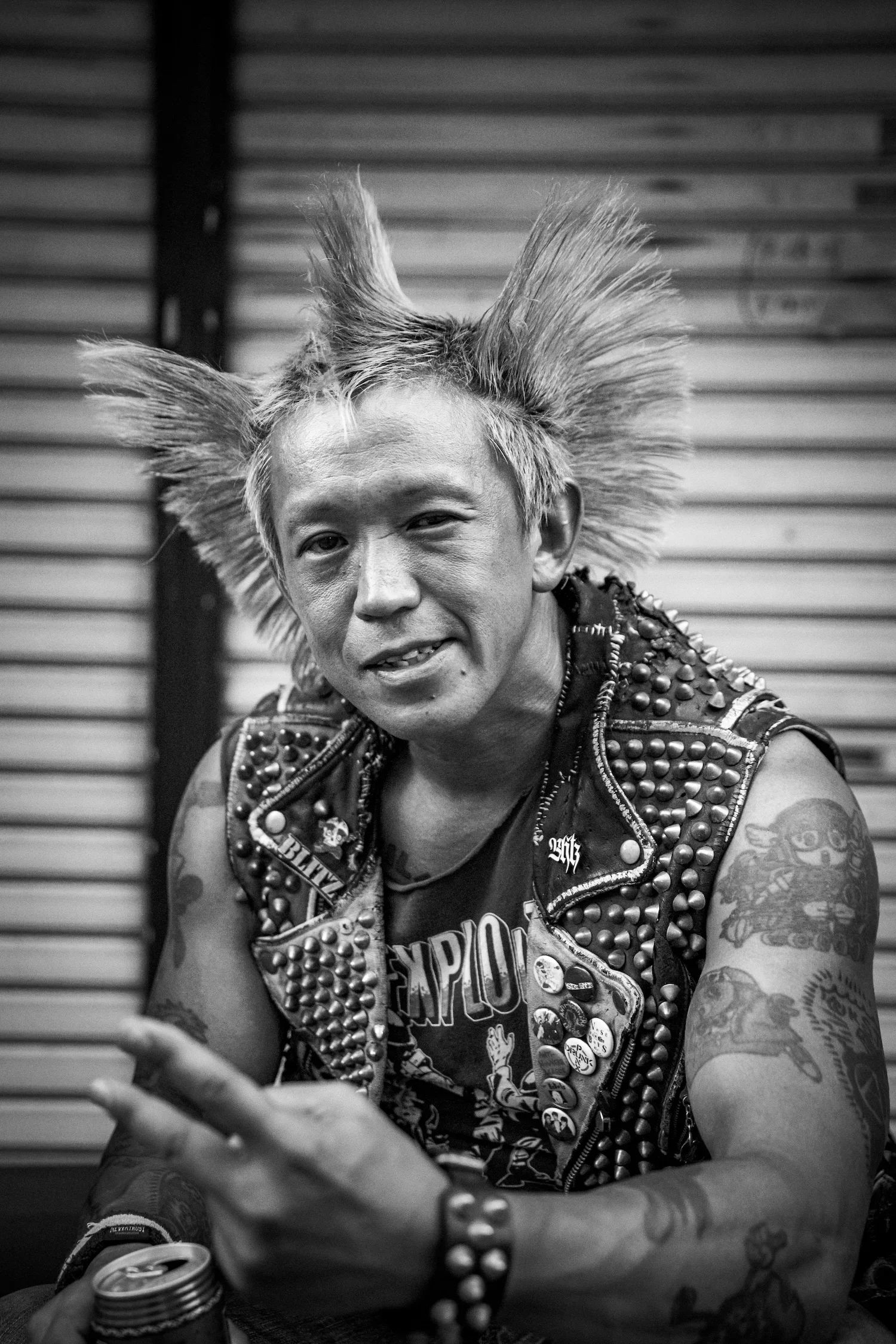 Black and white photo of a man with spiky, voluminous hair, wearing a studded leather vest with patches and pins, and displaying tattoos on his arms. He holds a soda can in one hand, and the background consists of horizontal metal siding.