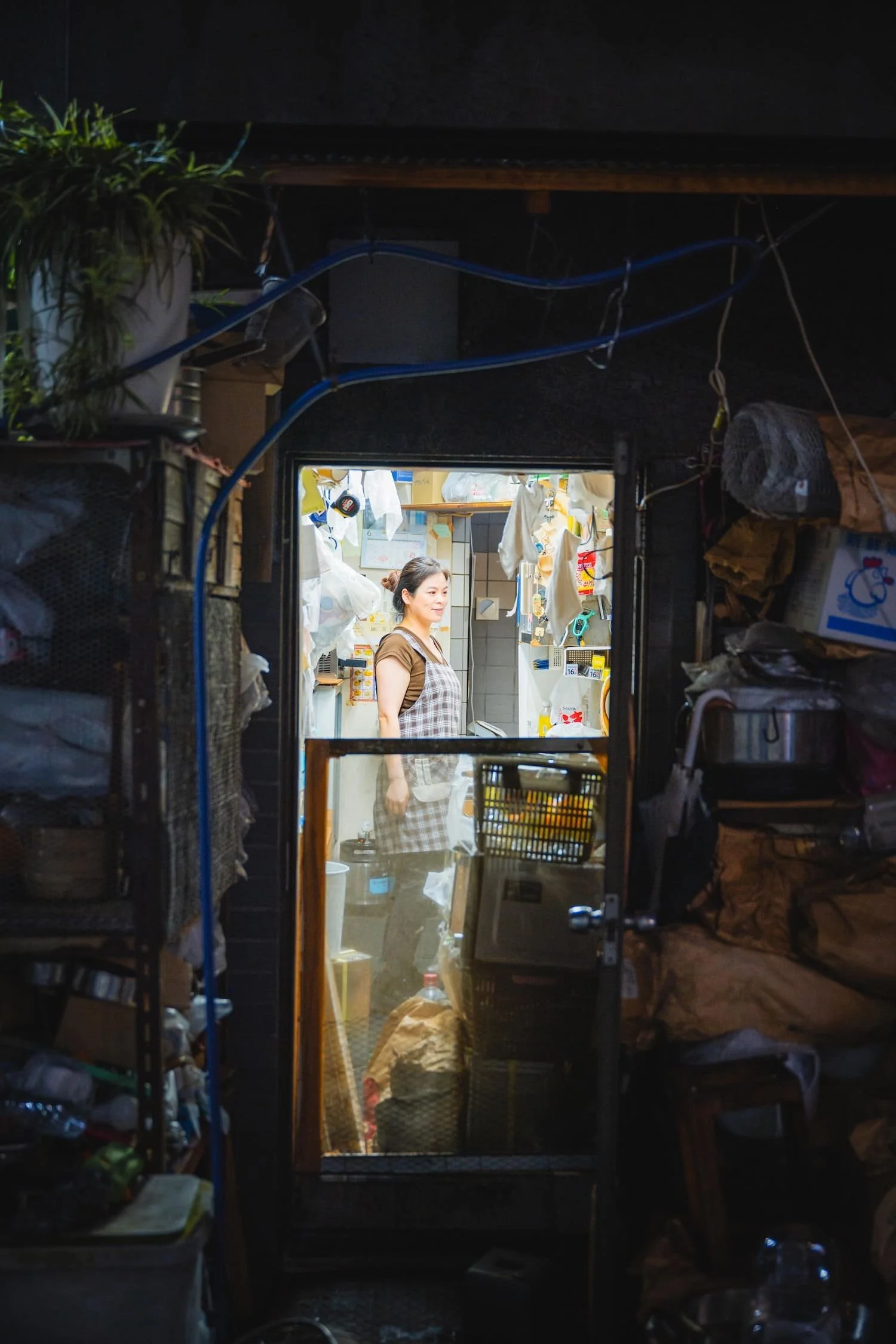 A woman standing inside a small, cluttered kitchen viewed through a glass door, with various hanging items and shelves behind her.